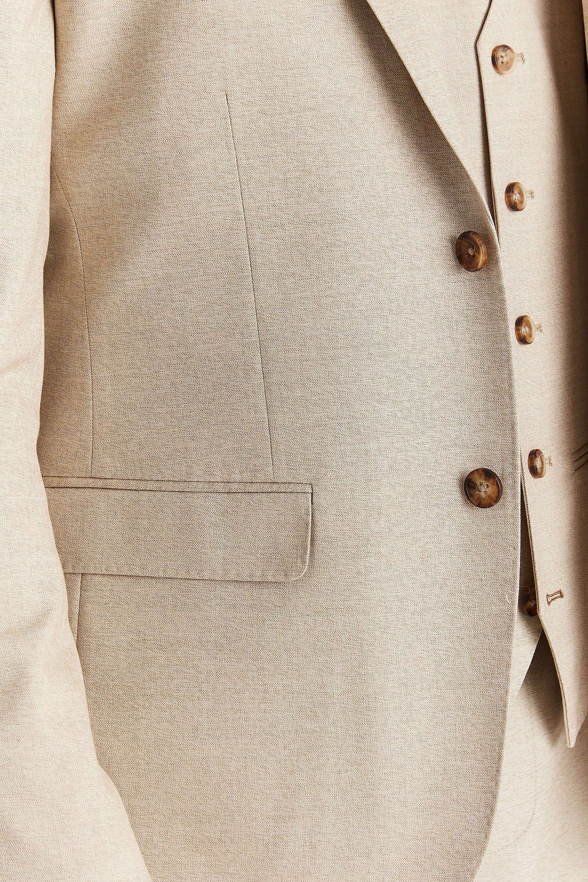Close-up view of the beige blazer showing the fine woven surface, brown buttons and a front flap pocket.