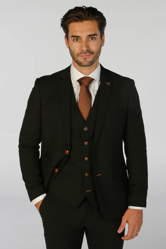 Model wearing a black textured three-piece suit with brown tie and pocket square, hands in pockets, shown from waist up.