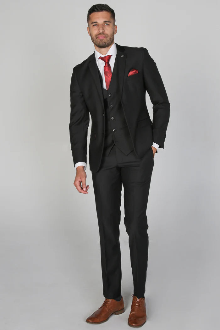 Full-body view of a model in a black three piece suit by Paul Andrew with red accessories and tan brogues.