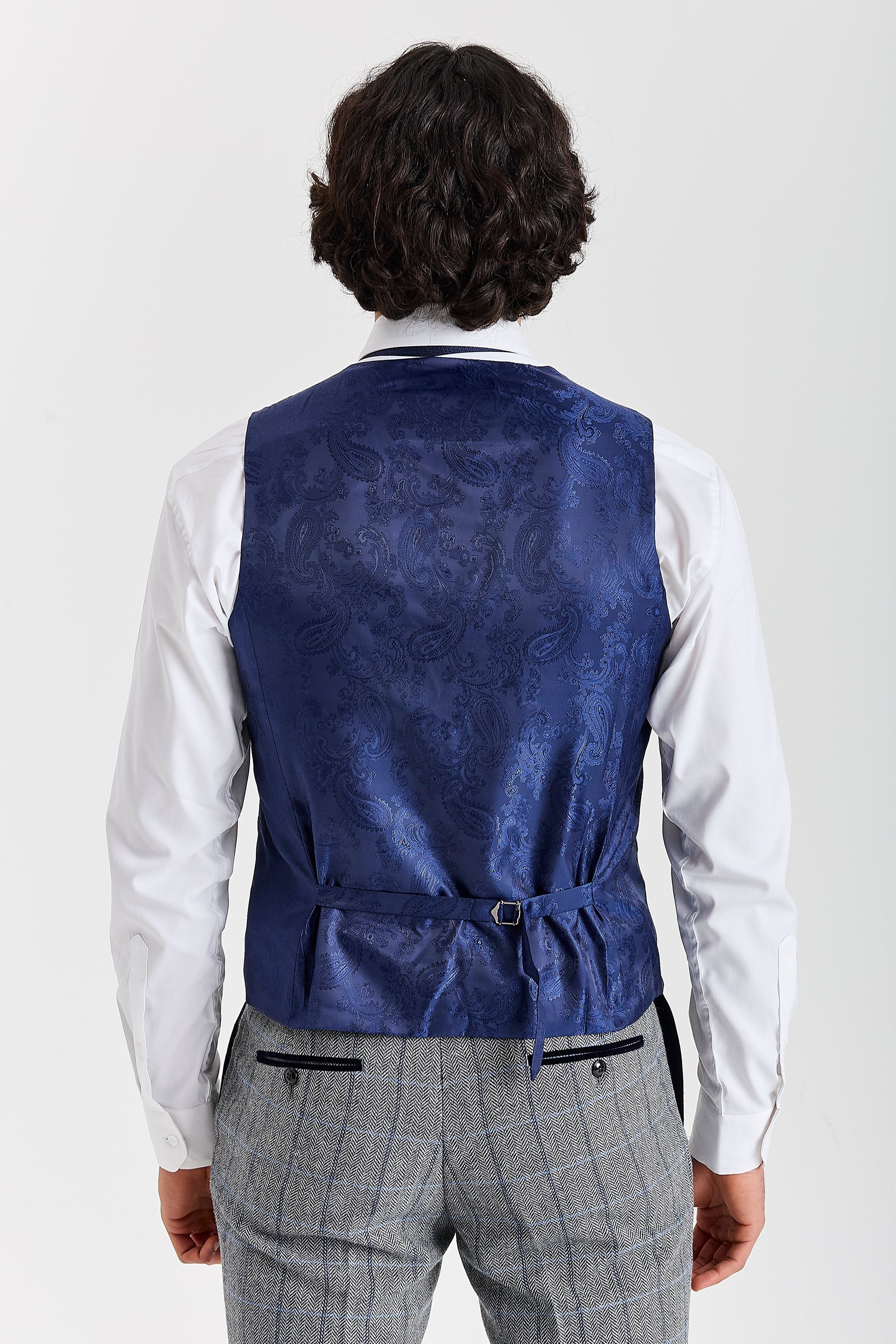 Rear view of a blue paisley waistcoat with adjustable strap, worn with a white shirt and grey herringbone tweed check trousers.