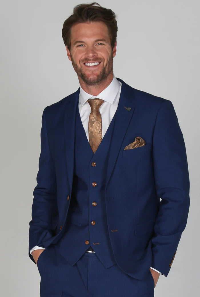 Close-up of a man in a textured blue three-piece suit with a gold paisley tie and matching pocket square, smiling confidently.