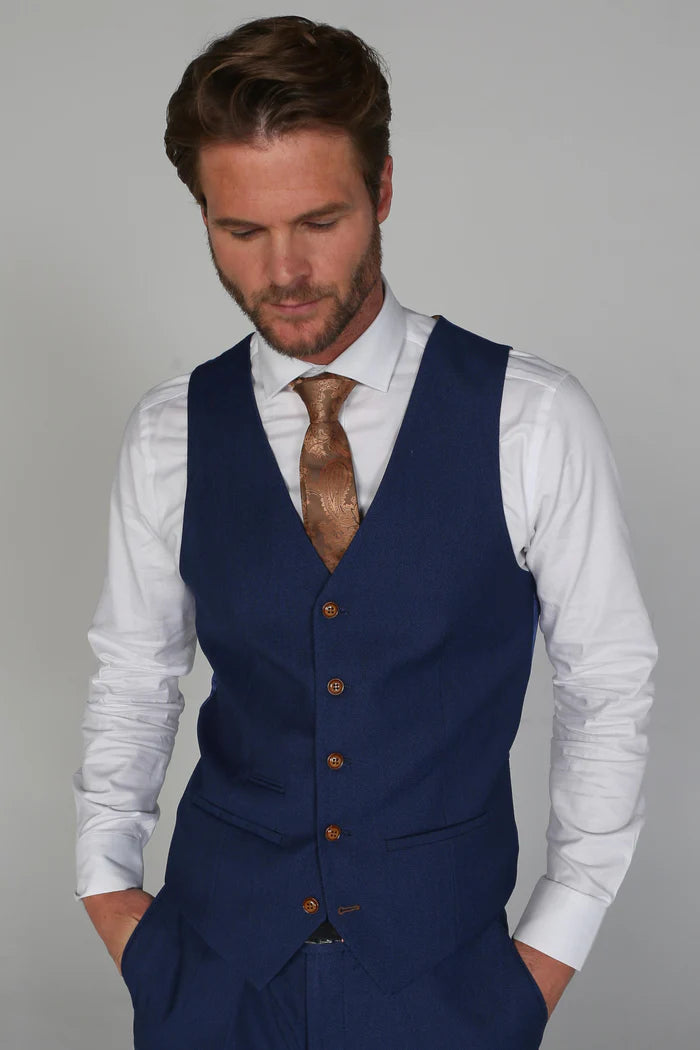 Man wearing the blue waistcoat with a goldish brown paisley tie and white shirt, hands in pockets, looking down slightly.