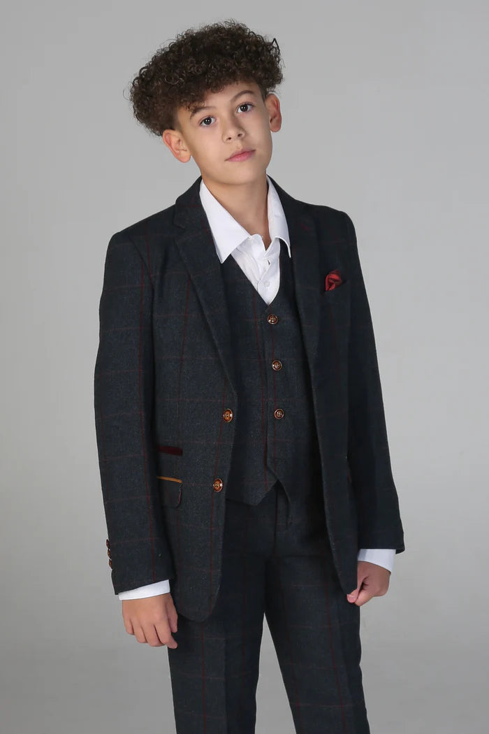 Boy in a navy three-piece suit with subtle check pattern, red pocket square, and brown buttons over white shirt.