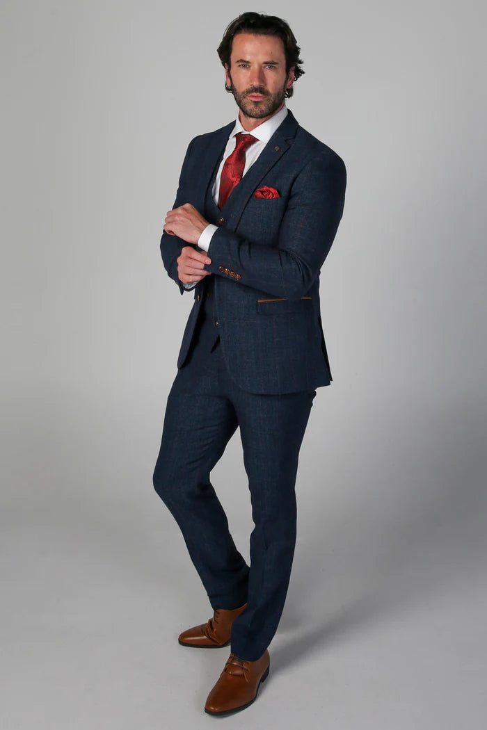 Full-body view of a man in a navy tweed check suit styled with a red tie and tan shoes, pointing to brown sleeve buttons.