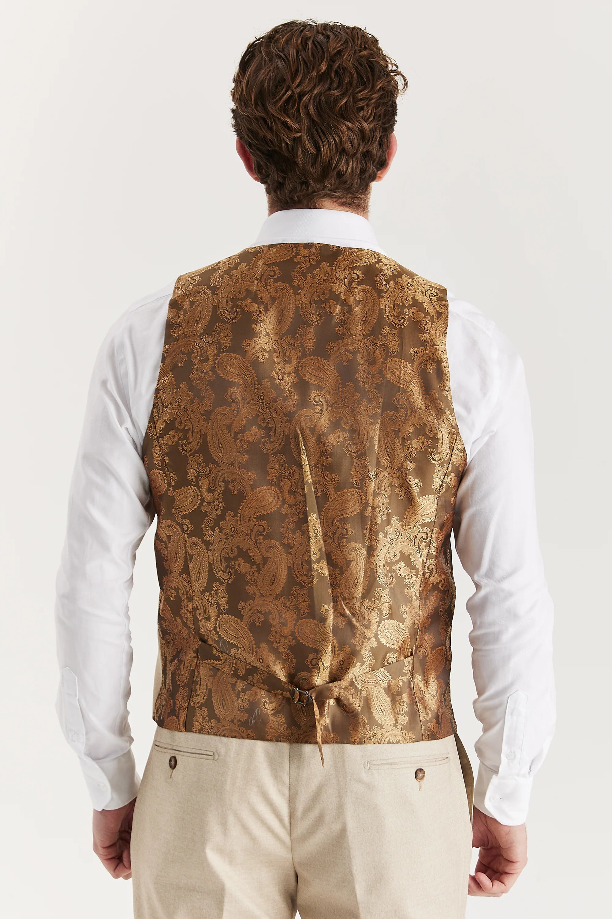 Rear view of the beige waistcoat with a brown paisley back panel and adjustable strap, worn with a white shirt and matching trousers.