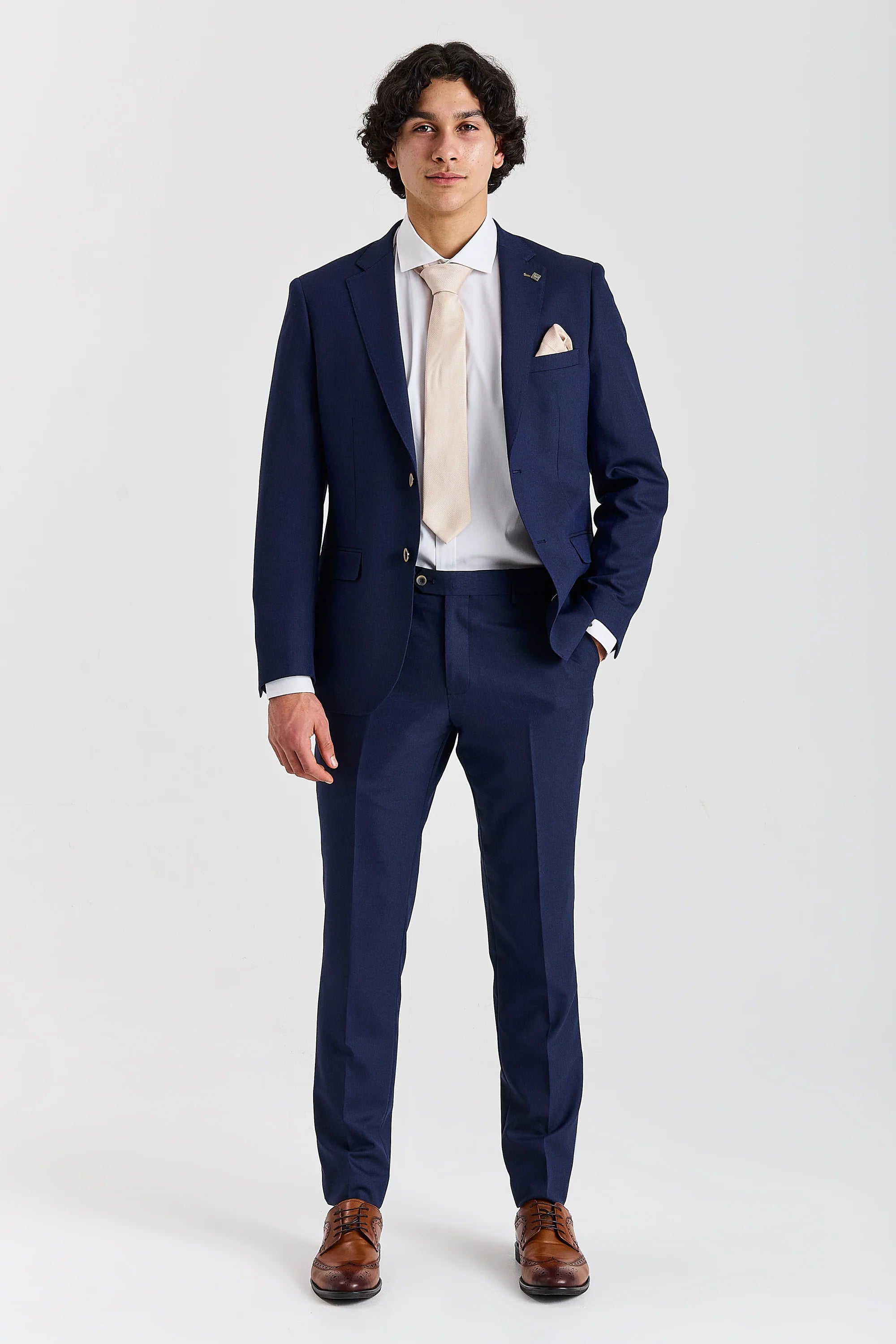Full-length front view of a man wearing a navy two-piece suit, paired with a light beige tie and brown brogues.