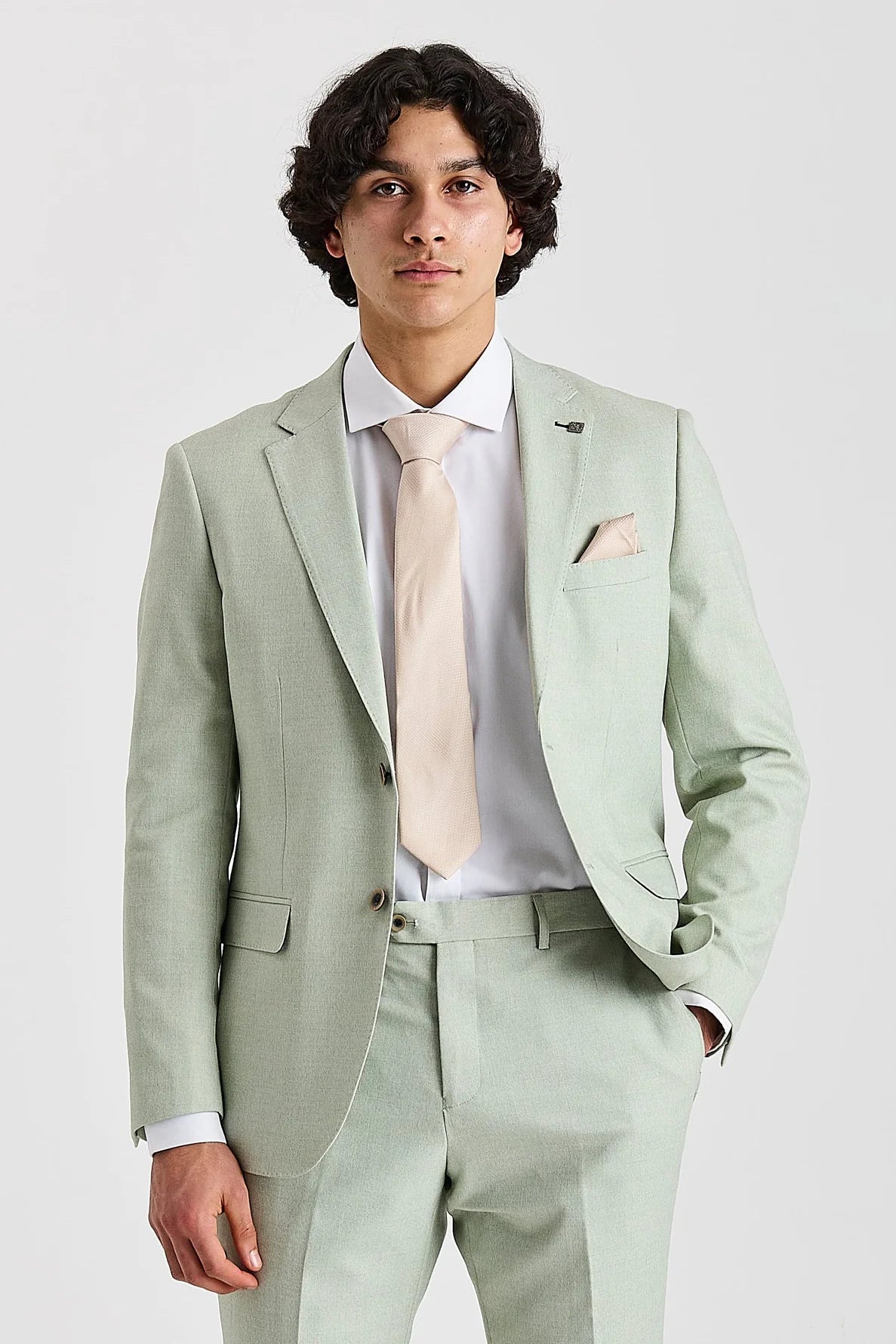 Waist-up front view of a man wearing a tailored light sage green suit jacket, styled with a crisp white shirt, light beige tie, and matching pocket square.