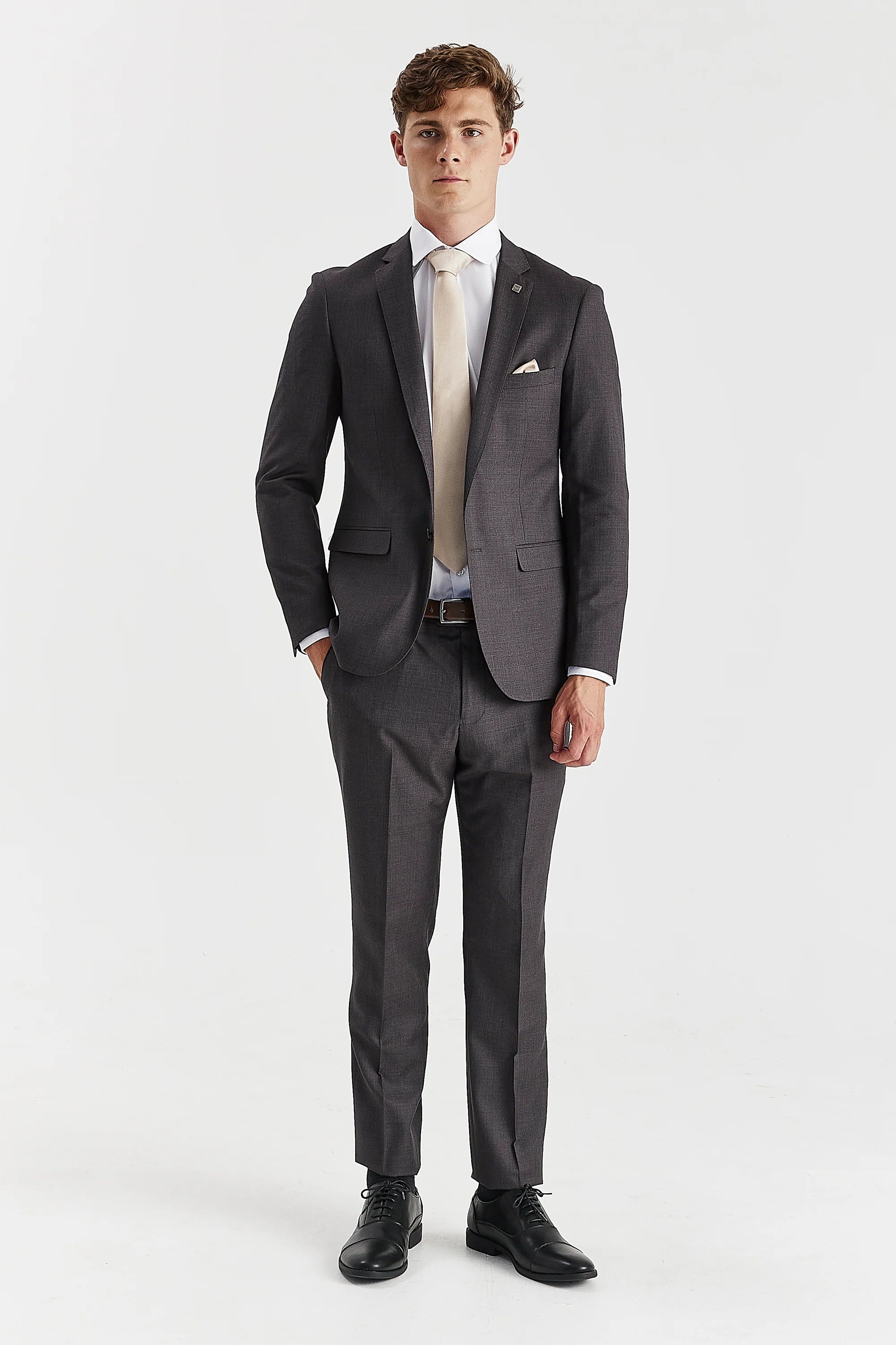 Full-length front view of a man wearing a charcoal grey two-piece suit, paired with a light beige tie and black leather dress shoes.