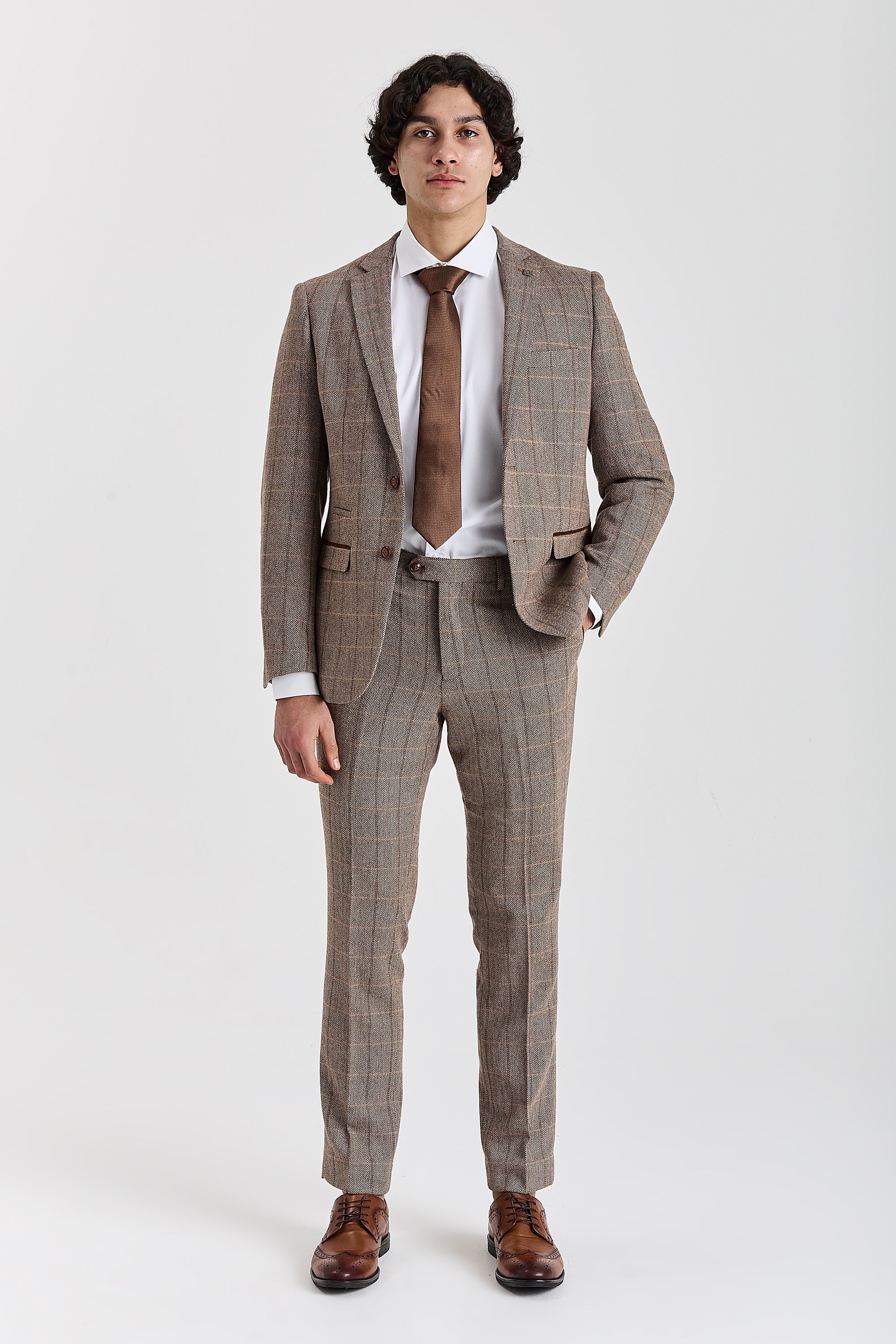 Full-length front view of a man modeling a brown herringbone tweed two-piece suit with a windowpane check pattern, paired with a white shirt, a brown tie, and brown brogue shoes
