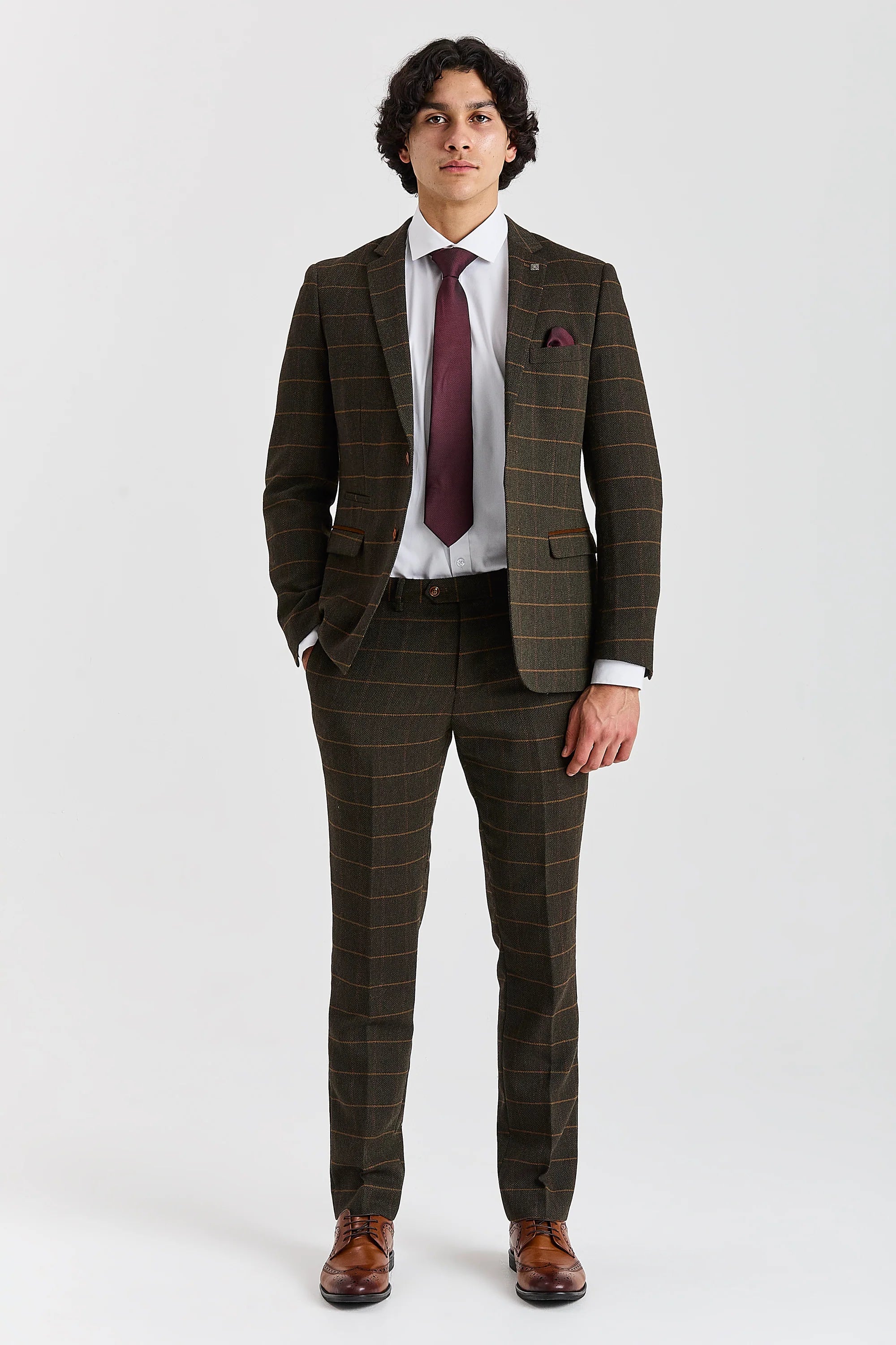 Full-length front view of a man modeling a green herringbone tweed two-piece suit with a windowpane check pattern, paired with a white shirt, burgundy tie, and brown leather brogue shoes.