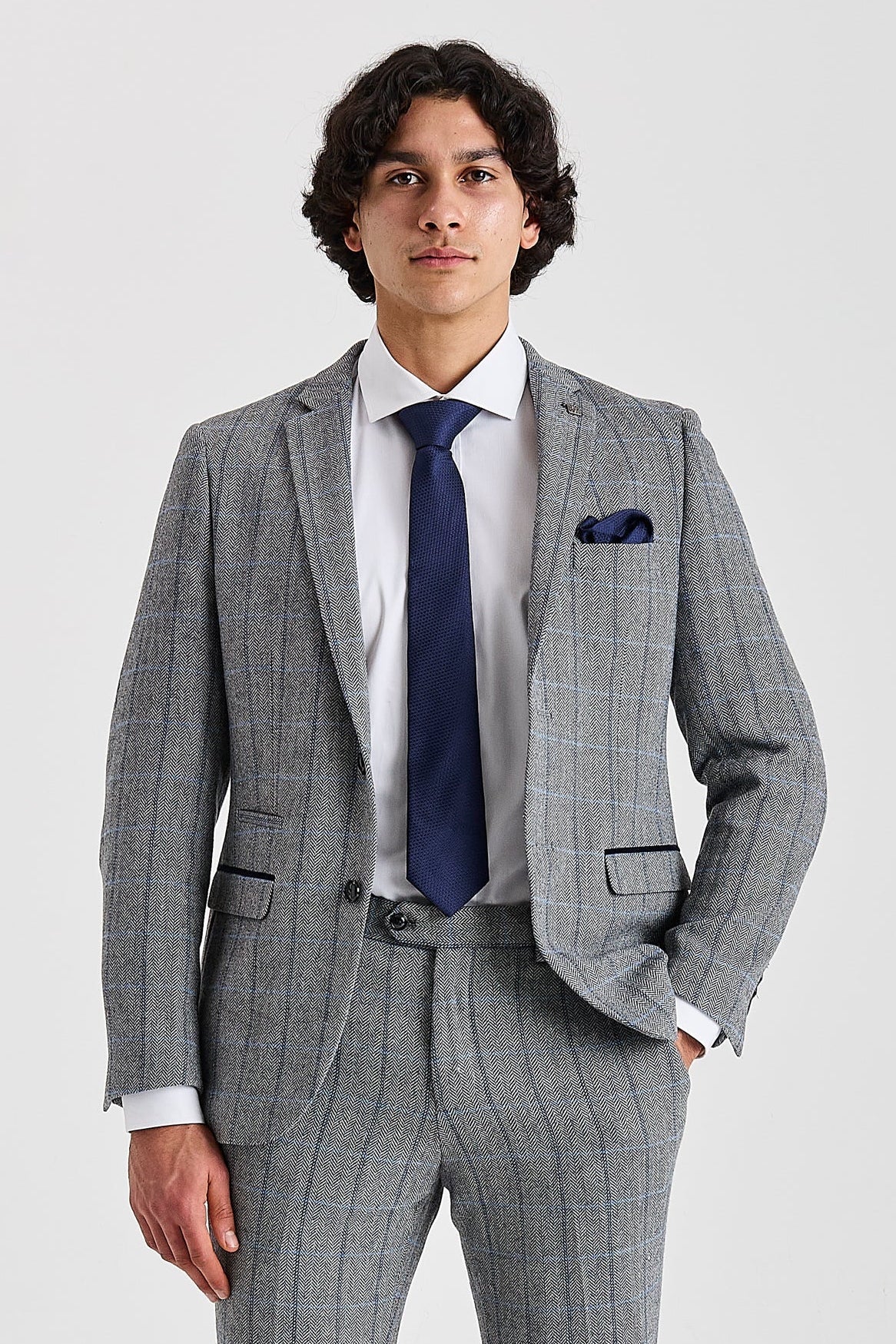 Knee-up view of a man wearing a grey herringbone tweed suit jacket with a windowpane check pattern. The jacket features subtle suede trimmings and is styled with a white shirt, navy tie, and navy pocket square