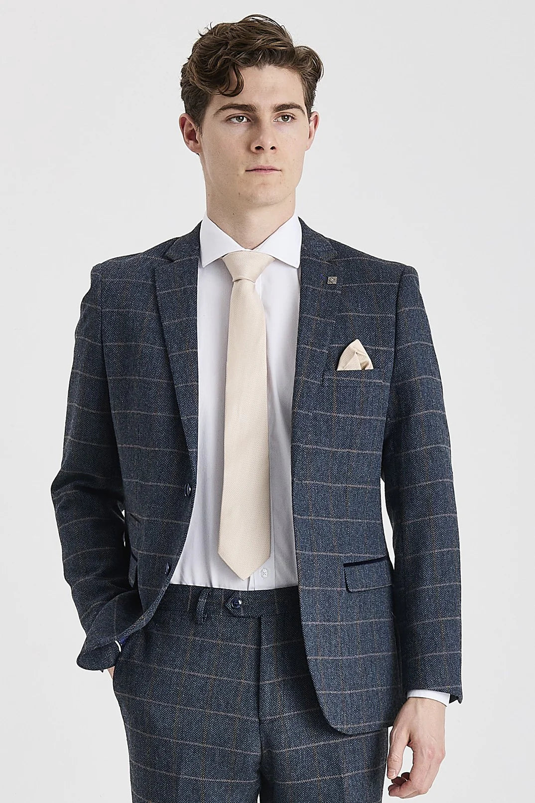 Knee-up view of a man wearing a navy herringbone tweed suit jacket with a windowpane check pattern. The jacket features subtle suede trimmings and is styled with a white shirt, a light beige tie, and a matching pocket square.