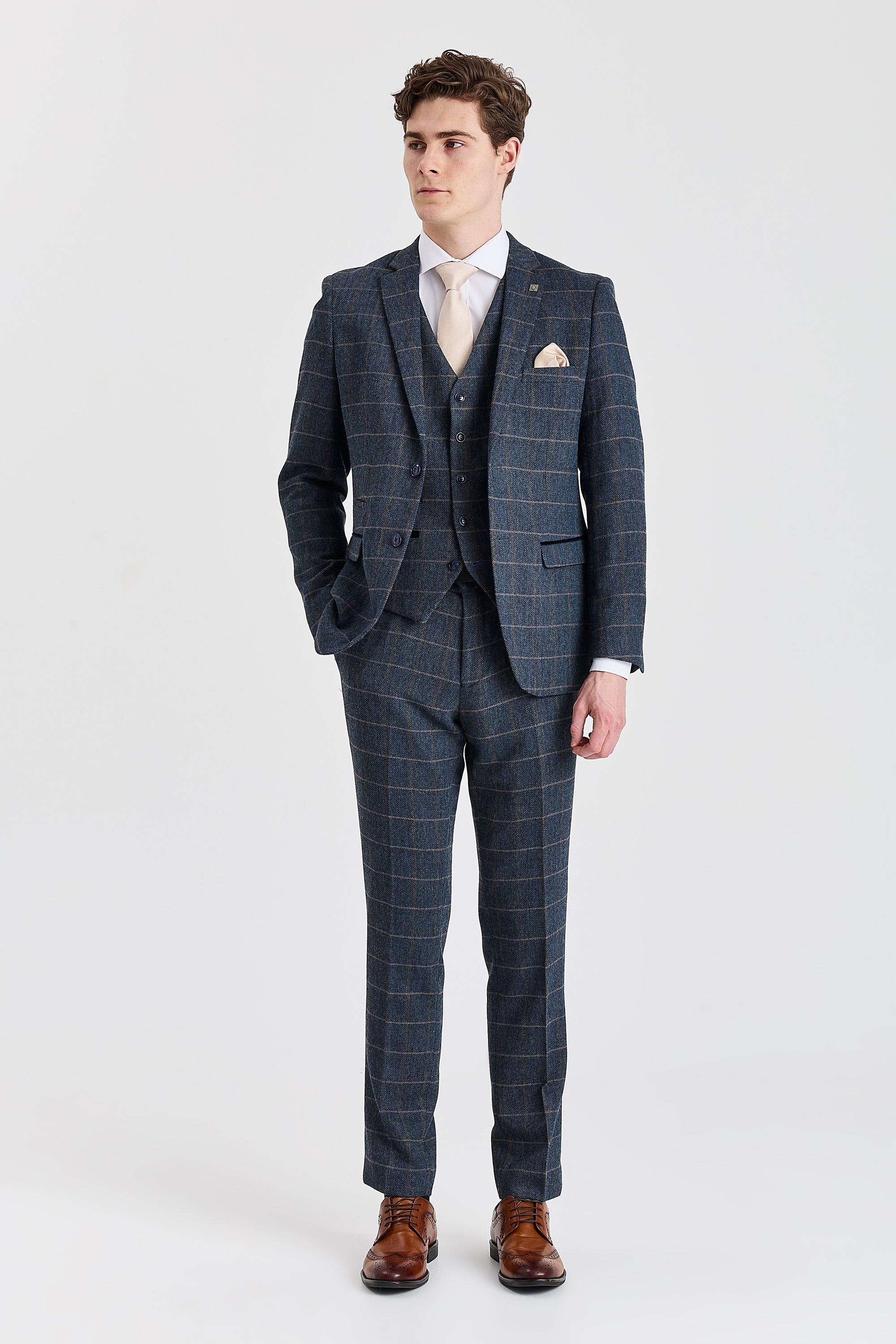 Full-length front view of a man wearing a Harris navy herringbone tweed three piece suit with a tan windowpane check, cream tie and brown brogue shoes.