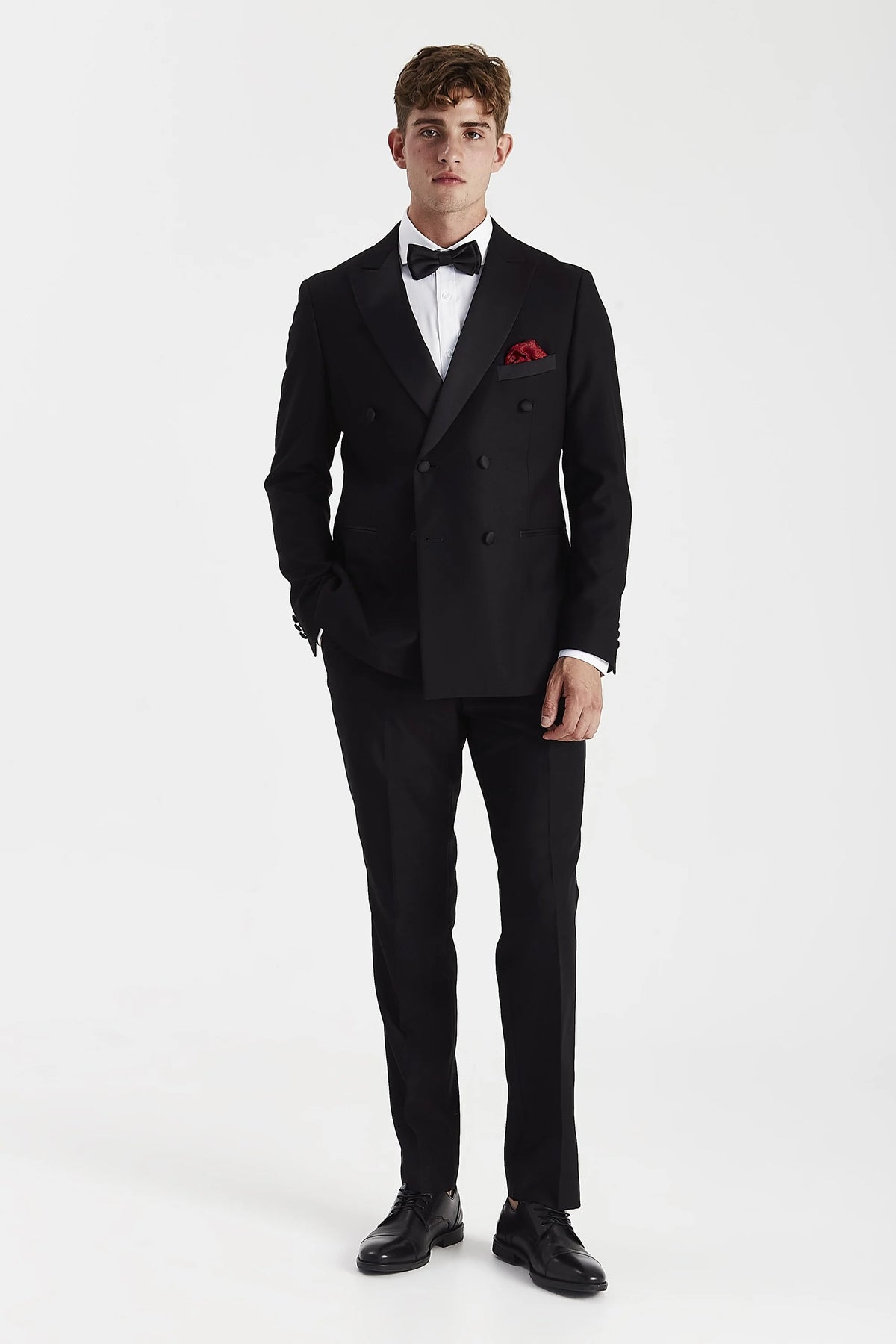 A man modeling a black double-breasted tuxedo with a white dress shirt, black bow tie, and a red pocket square, standing against a plain white studio background.