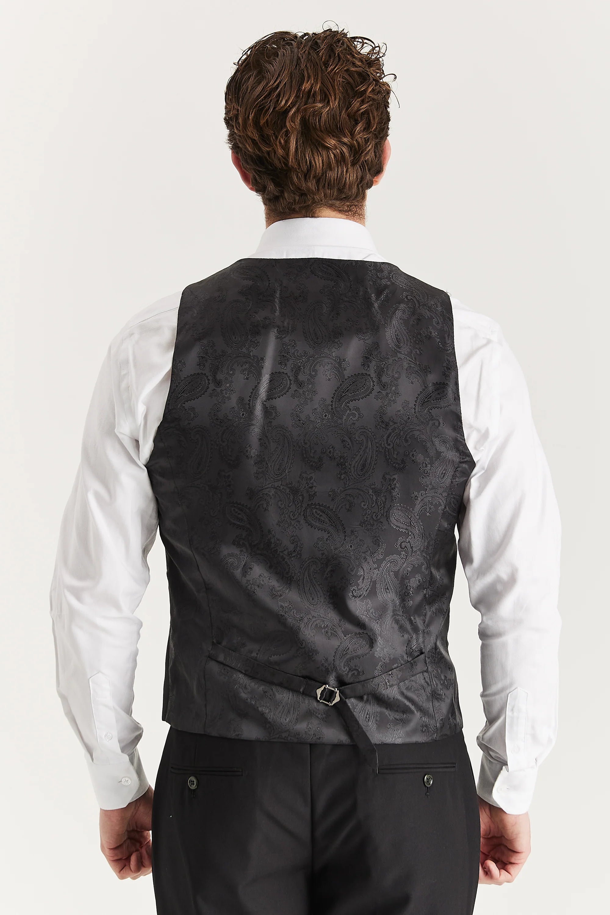 Rear view of the black waistcoat with a black paisley back panel and rear adjuster strap, worn with a white shirt and black trousers.