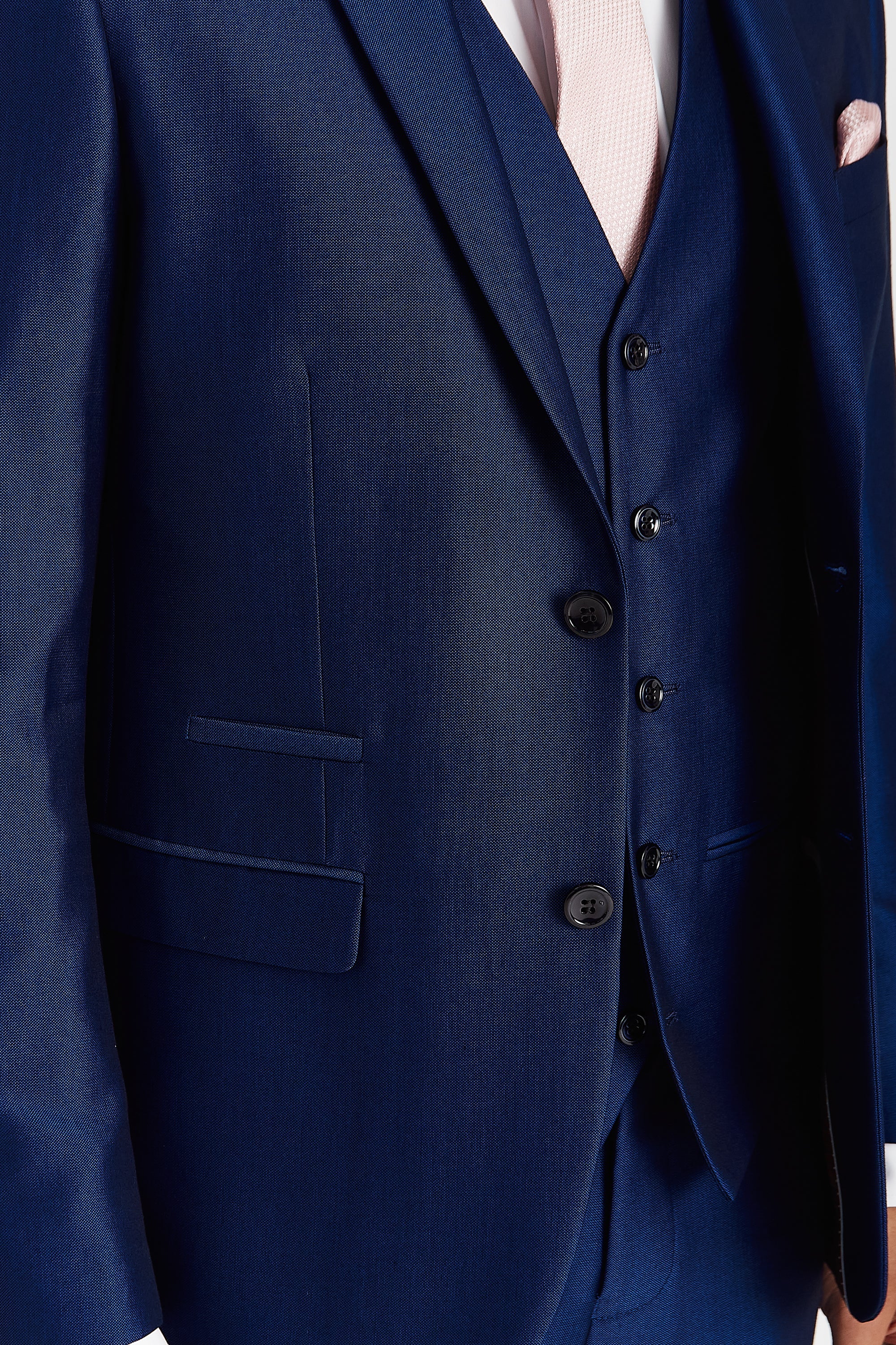 Detail view of blue suit fabric, showing the jacket, waistcoat, dark buttons, pocket trim, pink tie and pocket square.