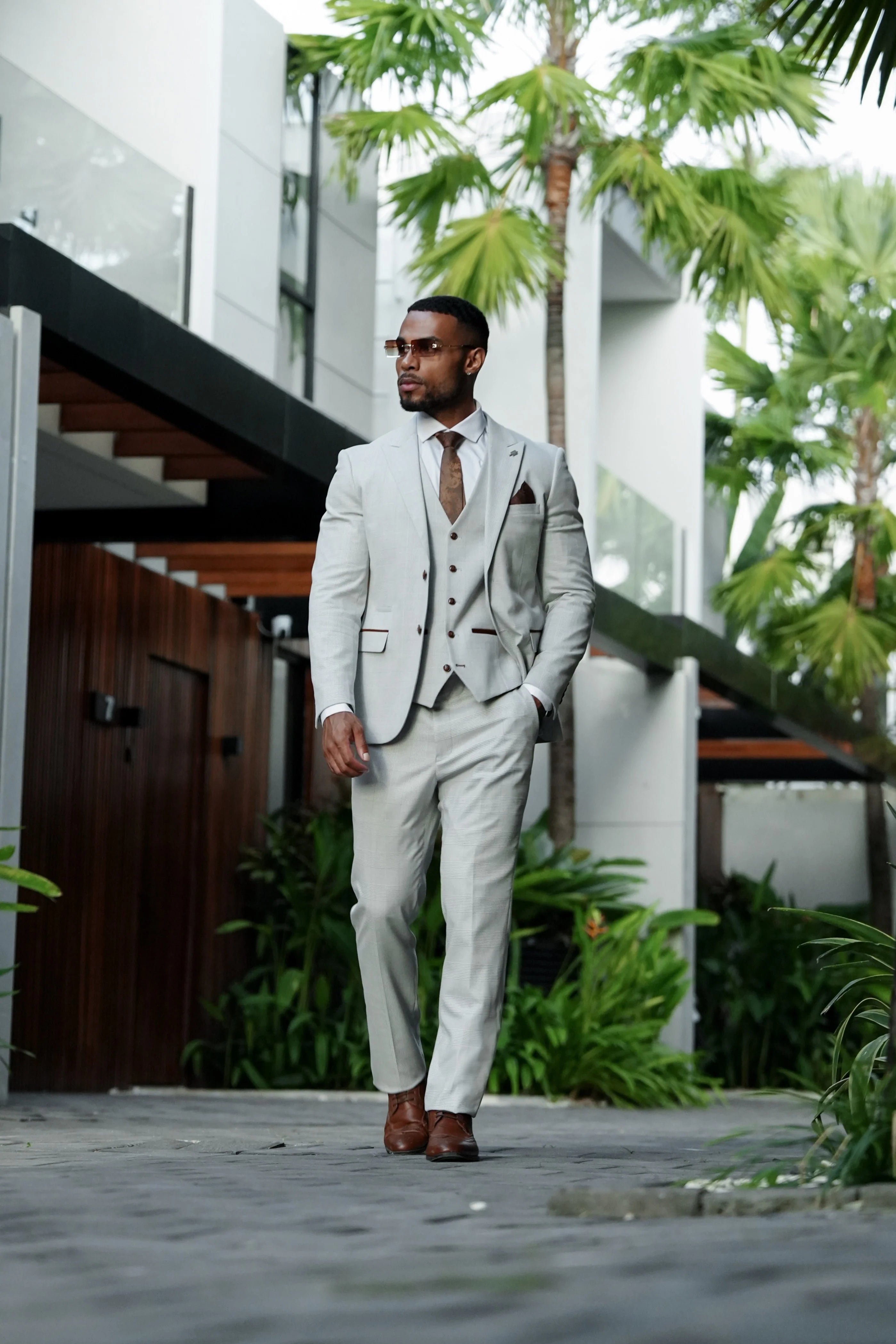 Full-length outdoor front view of a man wearing a stone three piece suit with a glen check pattern, white shirt, brown tie and brown shoes.