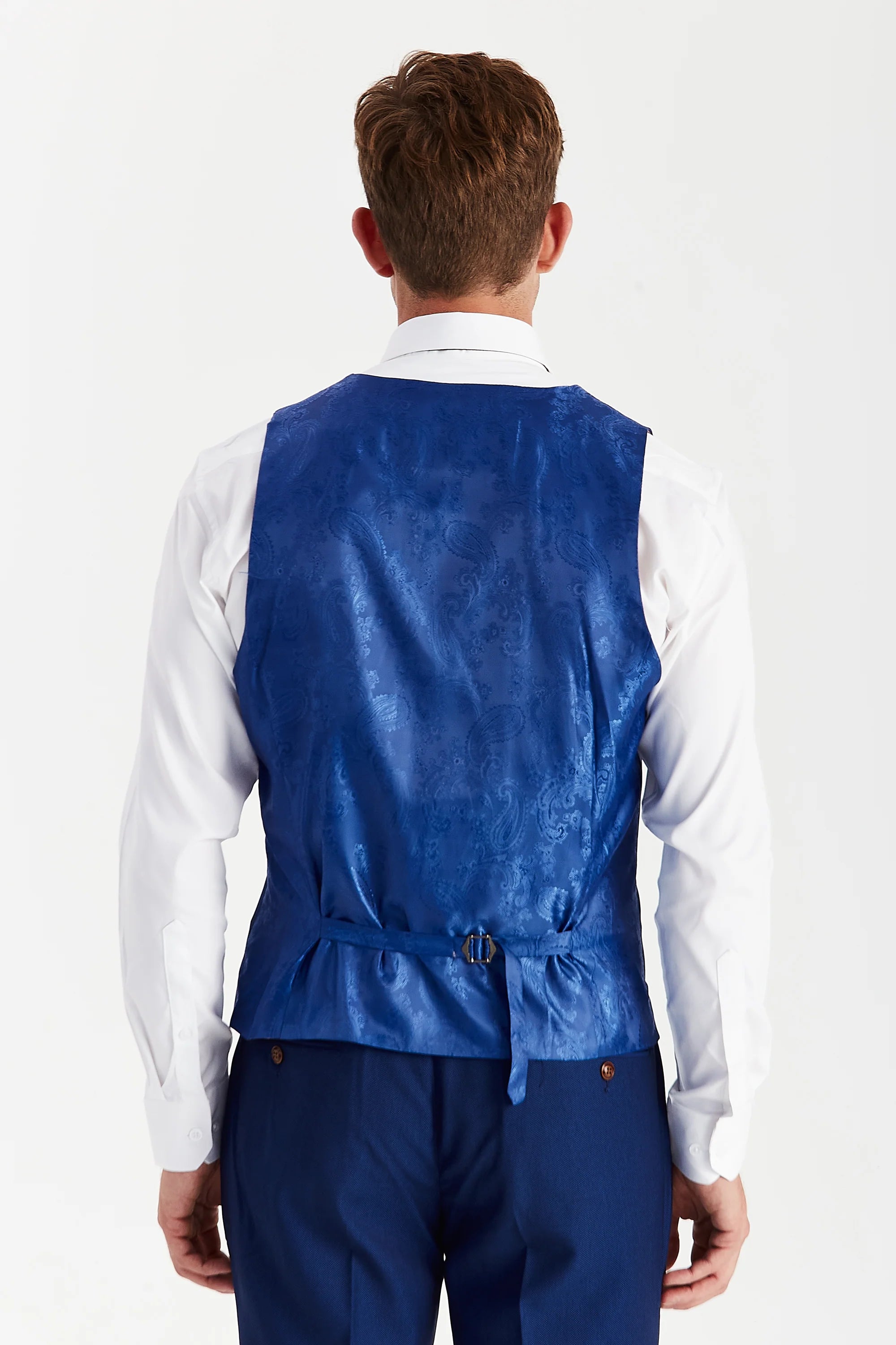 Back view of a blue paisley waistcoat panel with an adjustable strap, worn with blue trousers and a white shirt.