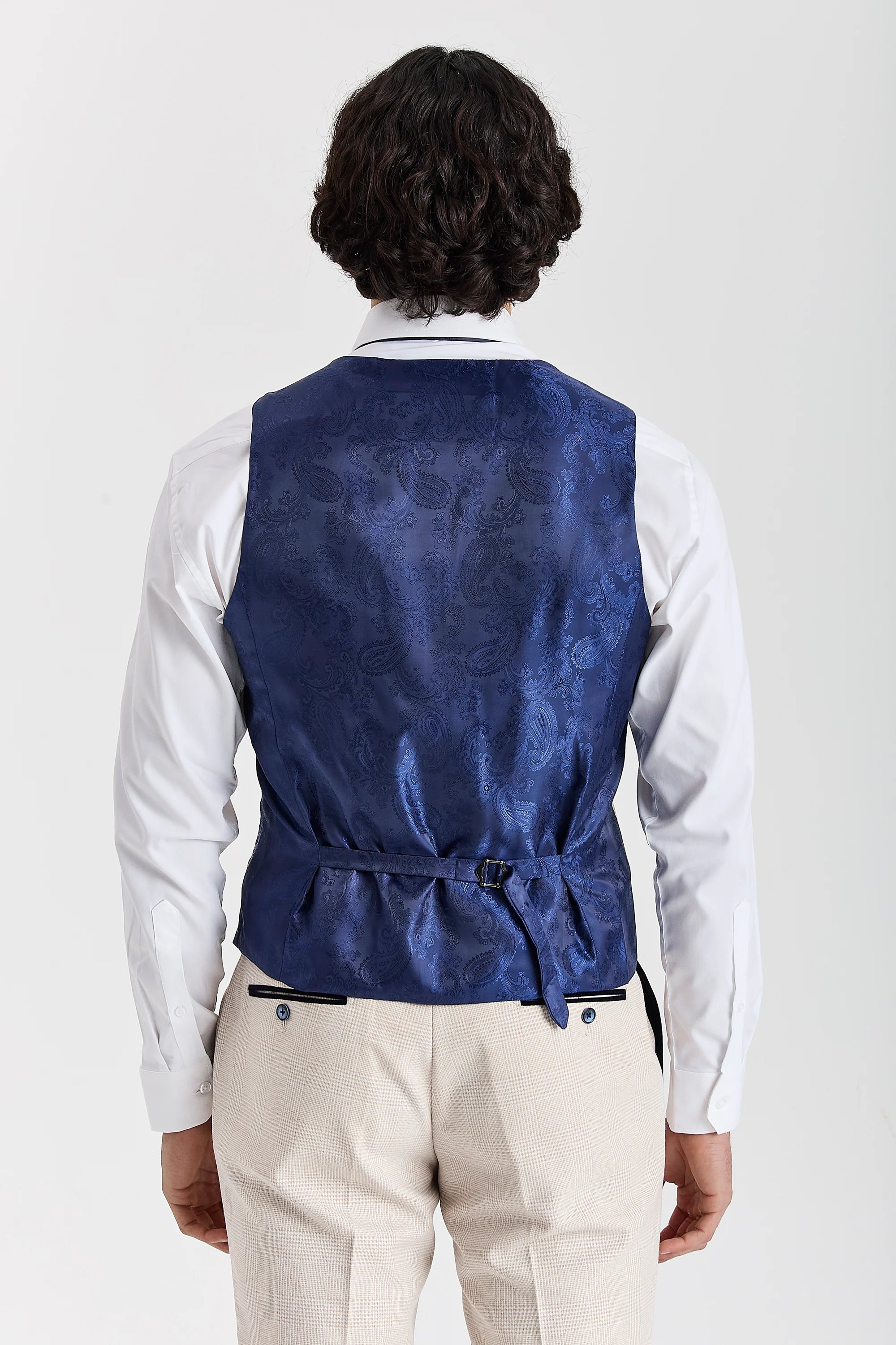 Rear view of the cream waistcoat with a blue paisley back panel and adjustable strap, worn with a white shirt and matching trousers.