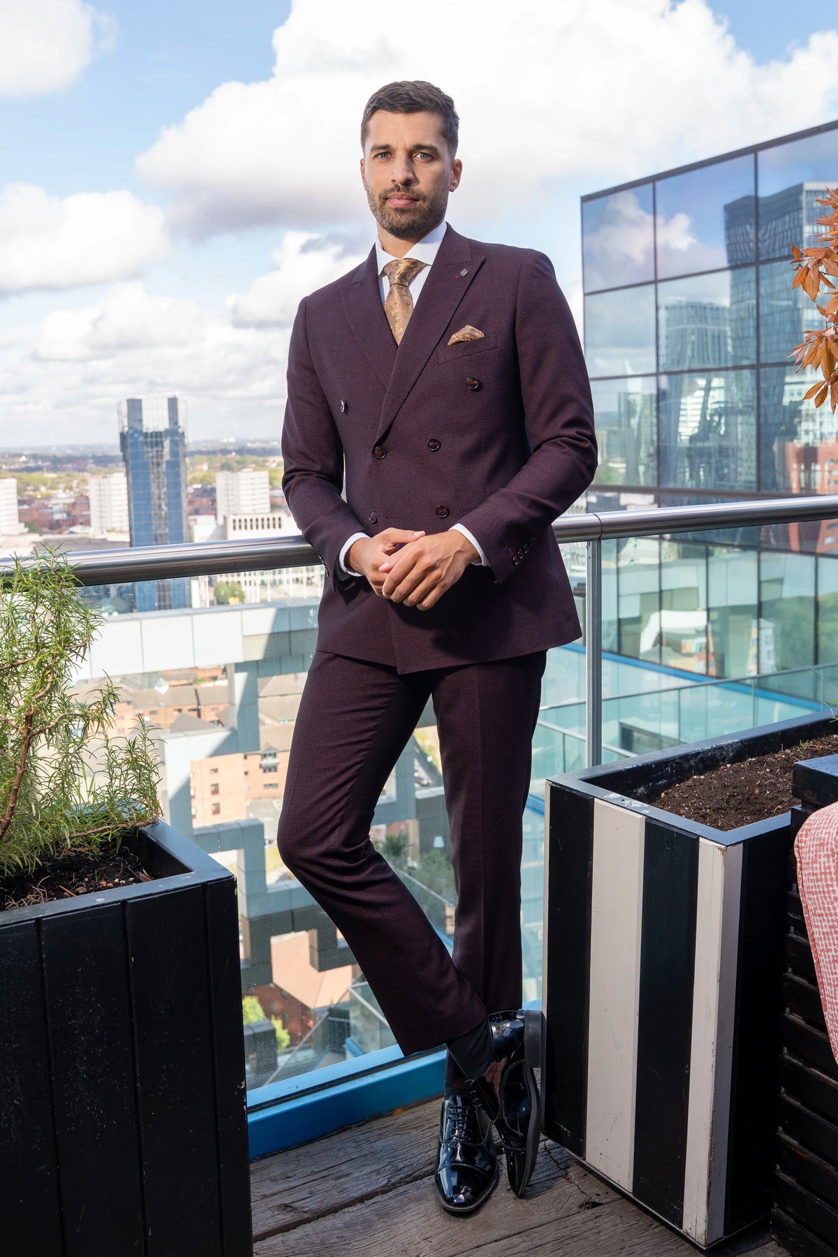 Model wearing the wine check suit on a rooftop terrace, styled with a white shirt, gold tie and matching pocket square.