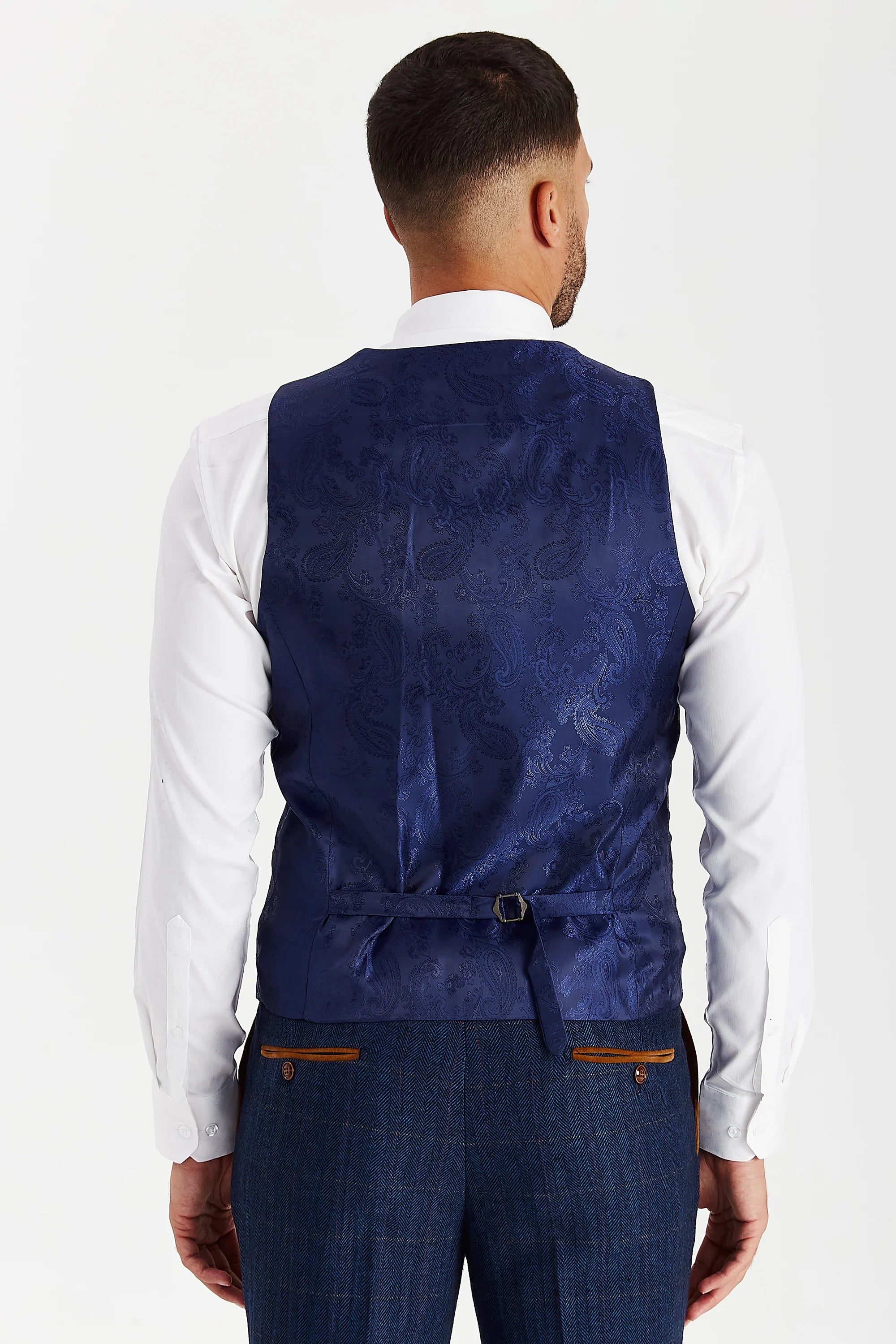 Rear view of the navy waistcoat with a blue paisley back panel and adjustable strap, worn with a white shirt and matching trousers.