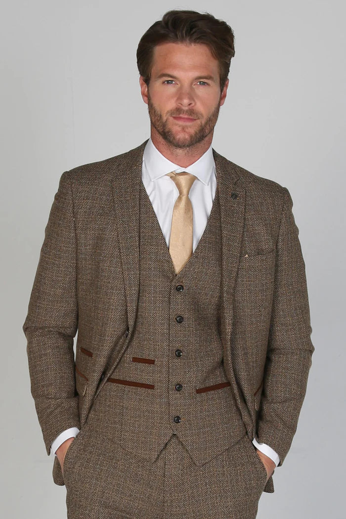 Close-up of model wearing Paul Andrew Ralph brown tweed three piece suit and cream tie with hands in pockets.