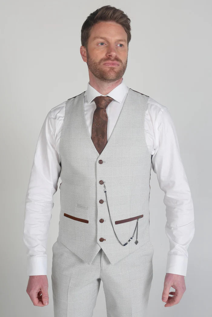 Model in a stone check waistcoat with a brown tie and chain accessory, hands relaxed at sides, showing full front styling.