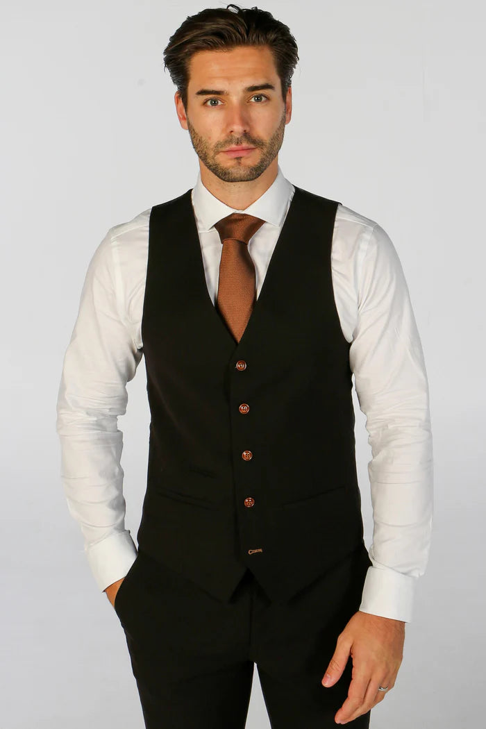 Man in a black waistcoat and white shirt with brown tie, posing with hands in pockets.