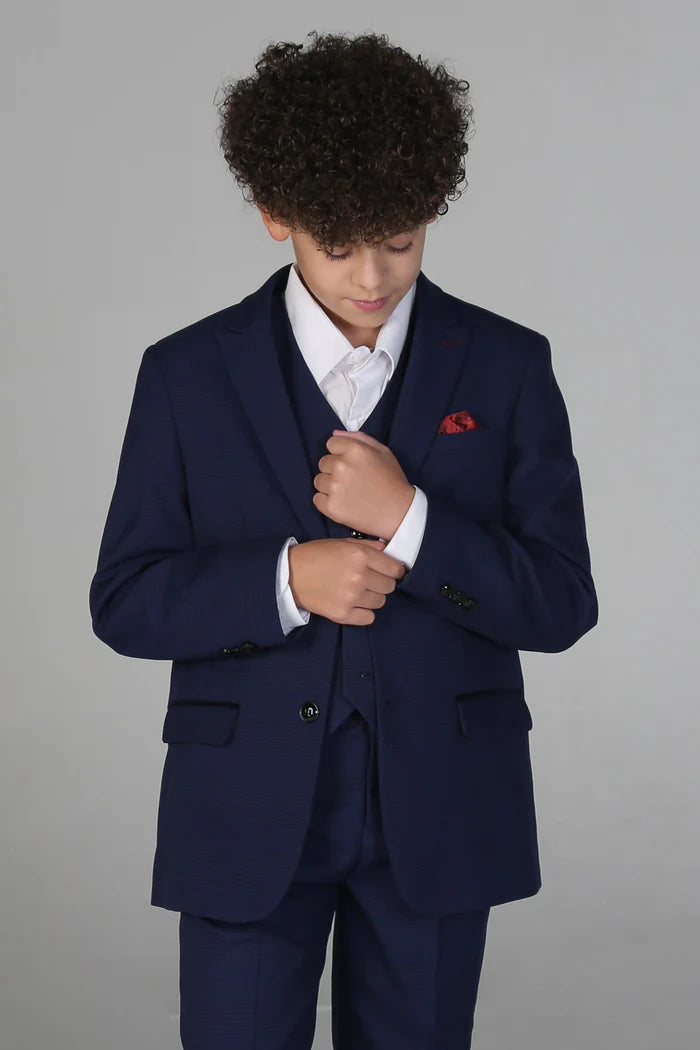 Boy adjusting navy suit jacket sleeve with red pocket square, showing formal three-piece outfit.