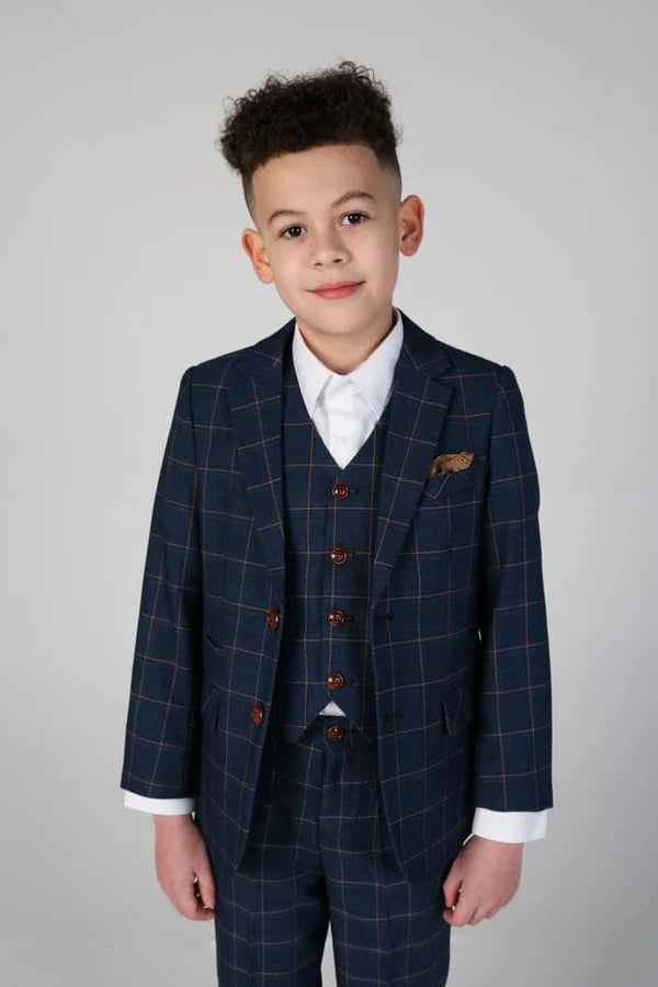 Young boy wearing navy blue windowpane check three-piece suit with white shirt and brown shoes, standing against light background.
