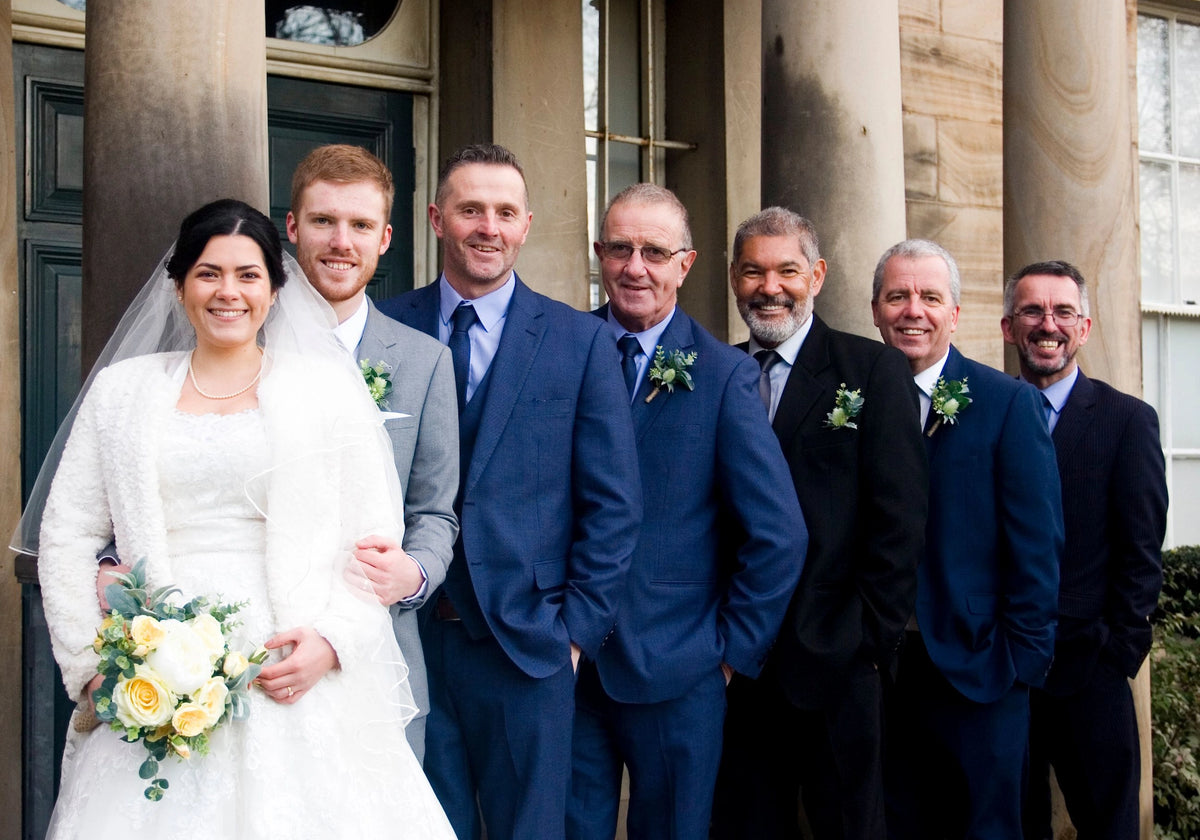 Bride and groom posing with groomsmen and wedding party wearing Navy and Black suits outside a wedding venue.