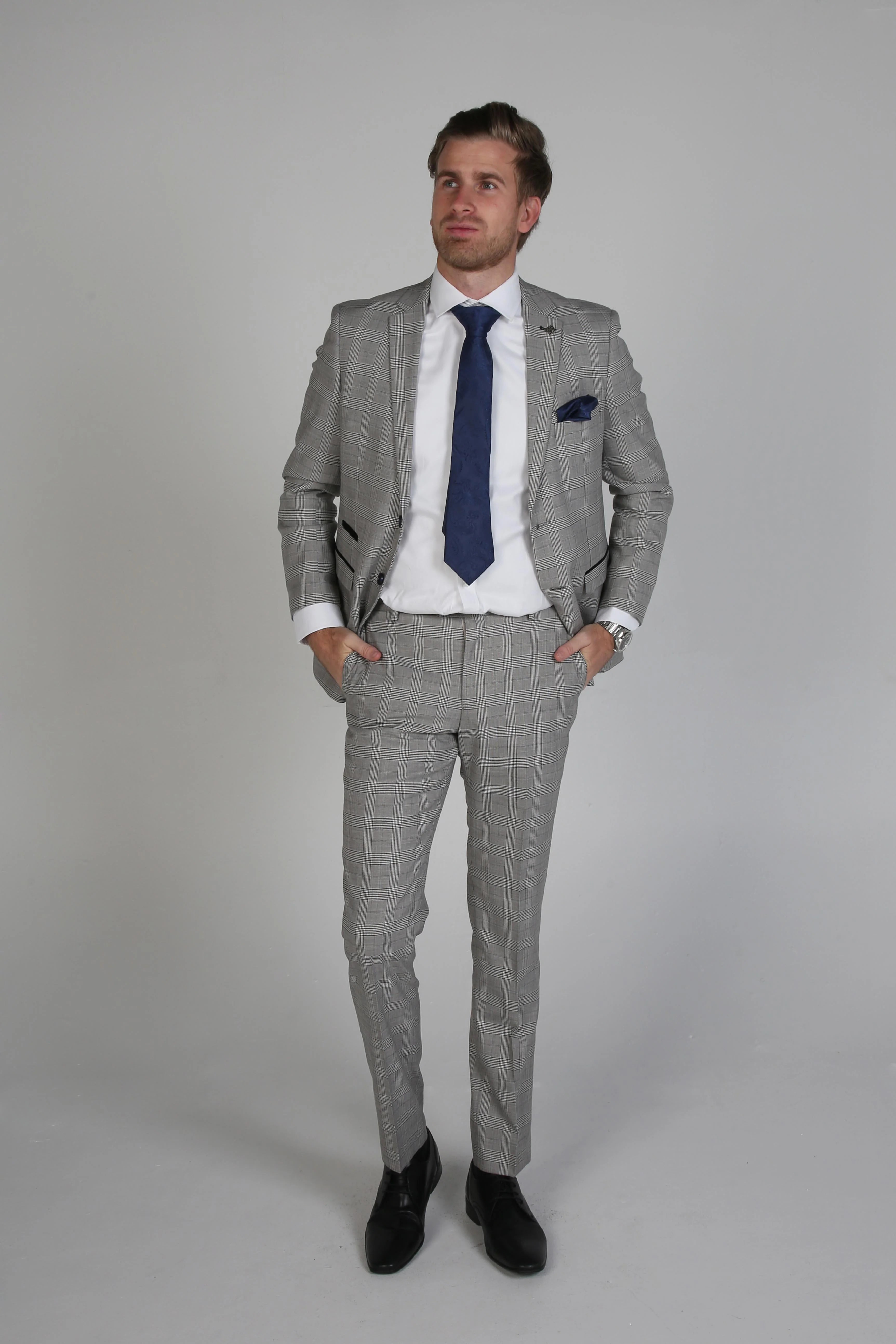 Man wearing a Hugo grey checkered three-piece suit with a white dress shirt and blue tie, looking up with hands in pockets.