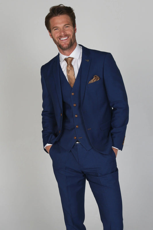 Man wearing a Mayfair, a plain dark blue three-piece suit with a white dress shirt and brown tie, smiling with hands in pockets.