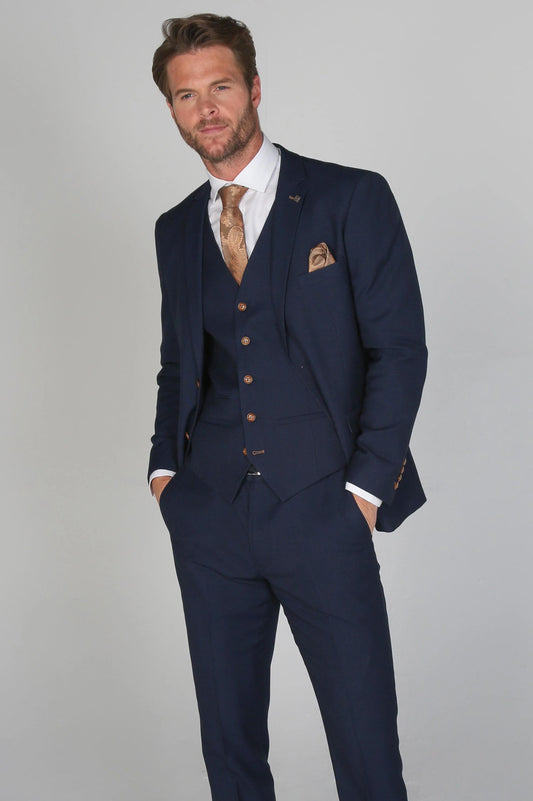 Mayfair Navy Men's three-piece suit with a white dress shirt and brown tie, model posing with hands in pockets.