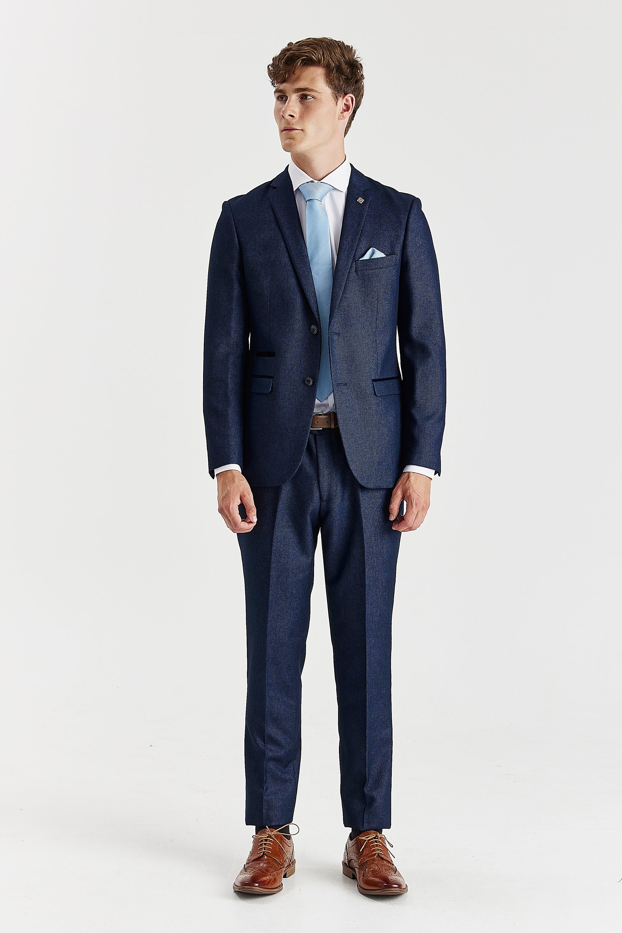 Full-length front view of a man wearing a tailored navy birdseye two-piece suit, styled with a light blue tie and brown brogues.