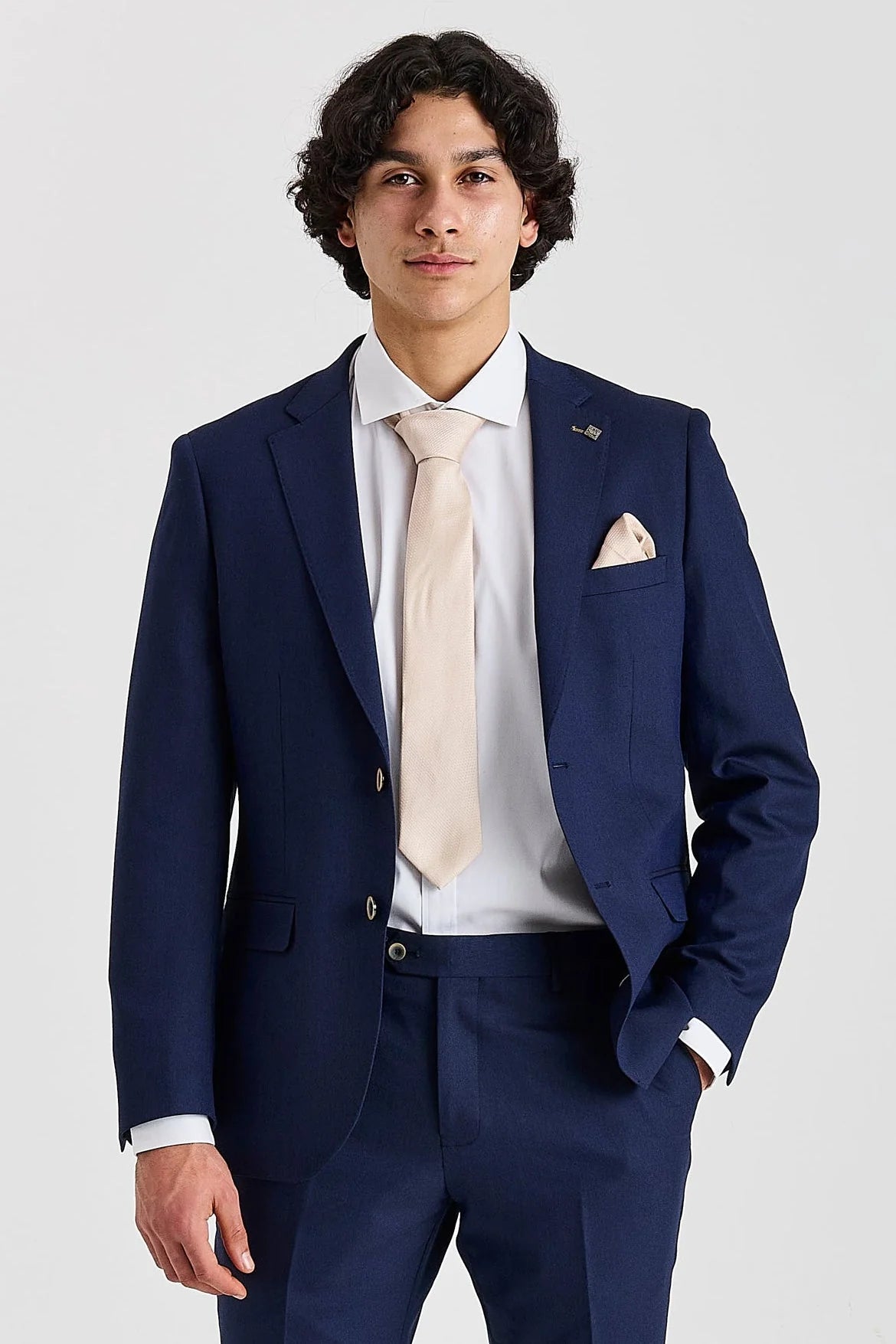 Waist-up front view of a man wearing a tailored navy men's suit jacket, styled with a white shirt, light beige tie, and matching pocket square.