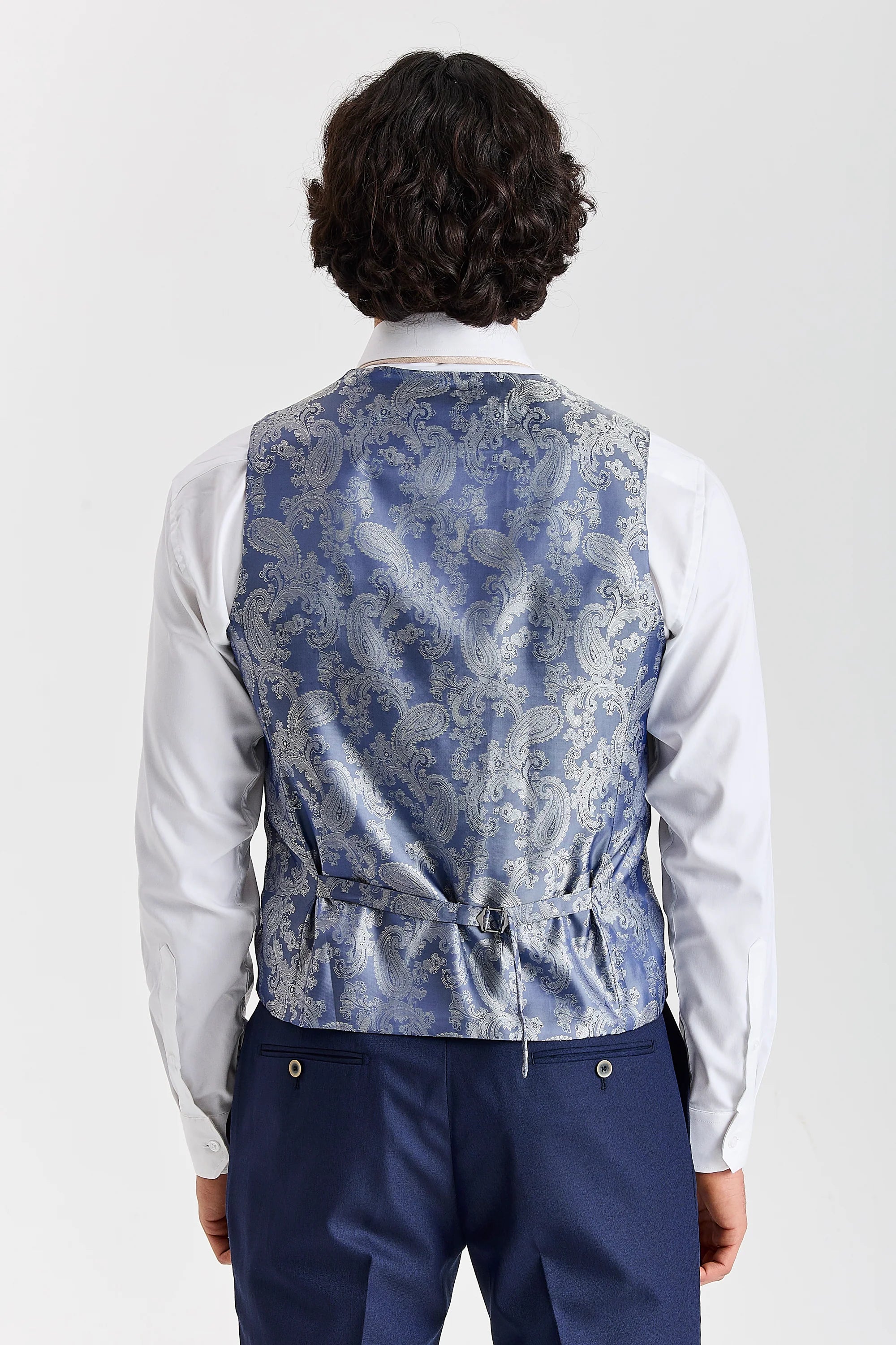 Rear view of the navy waistcoat with a blue paisley back panel and adjustable strap, worn with a white shirt and matching trousers.