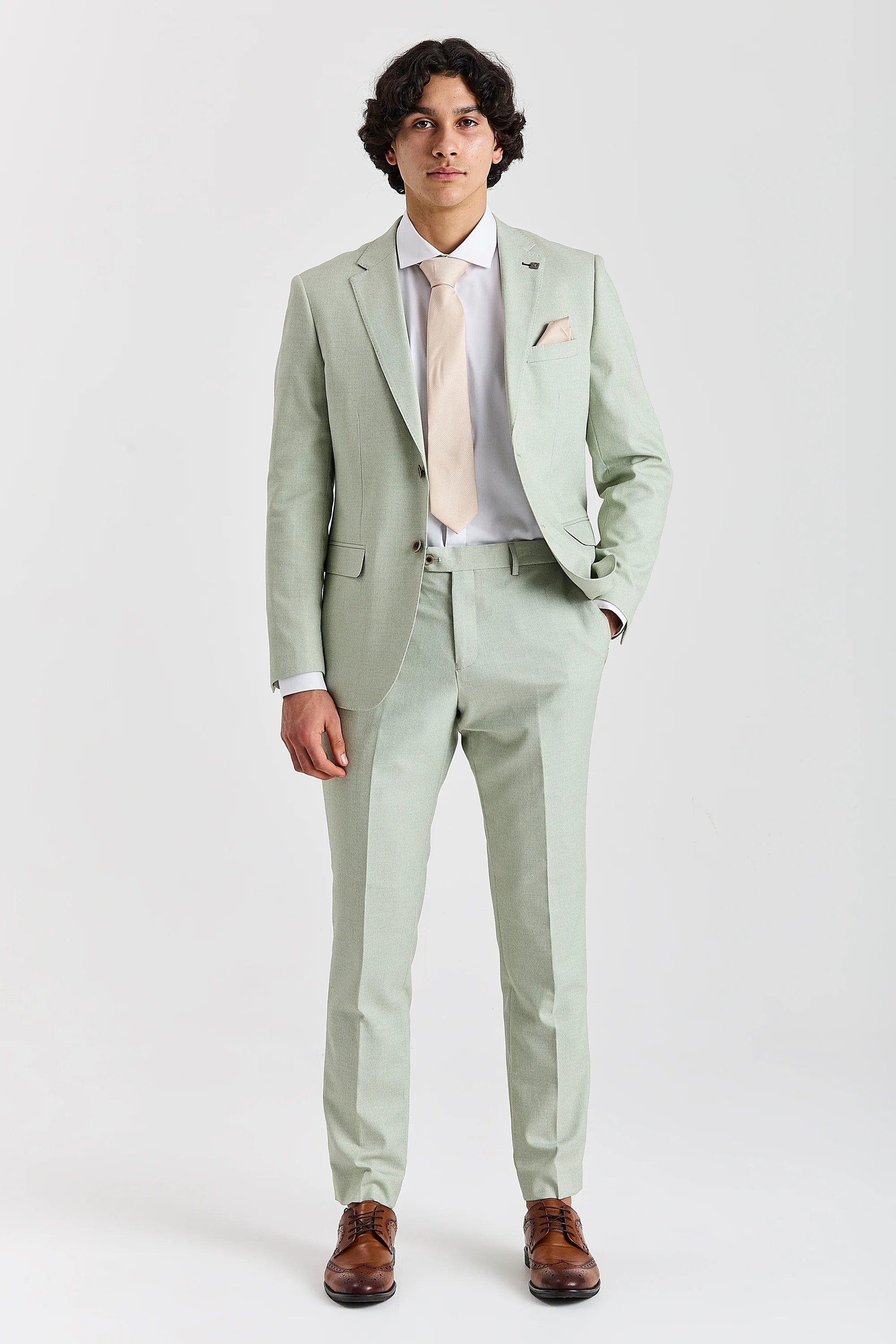 Full-length front view of a man modeling a light green two-piece suit, paired with a light beige tie and brown leather brogue shoes.