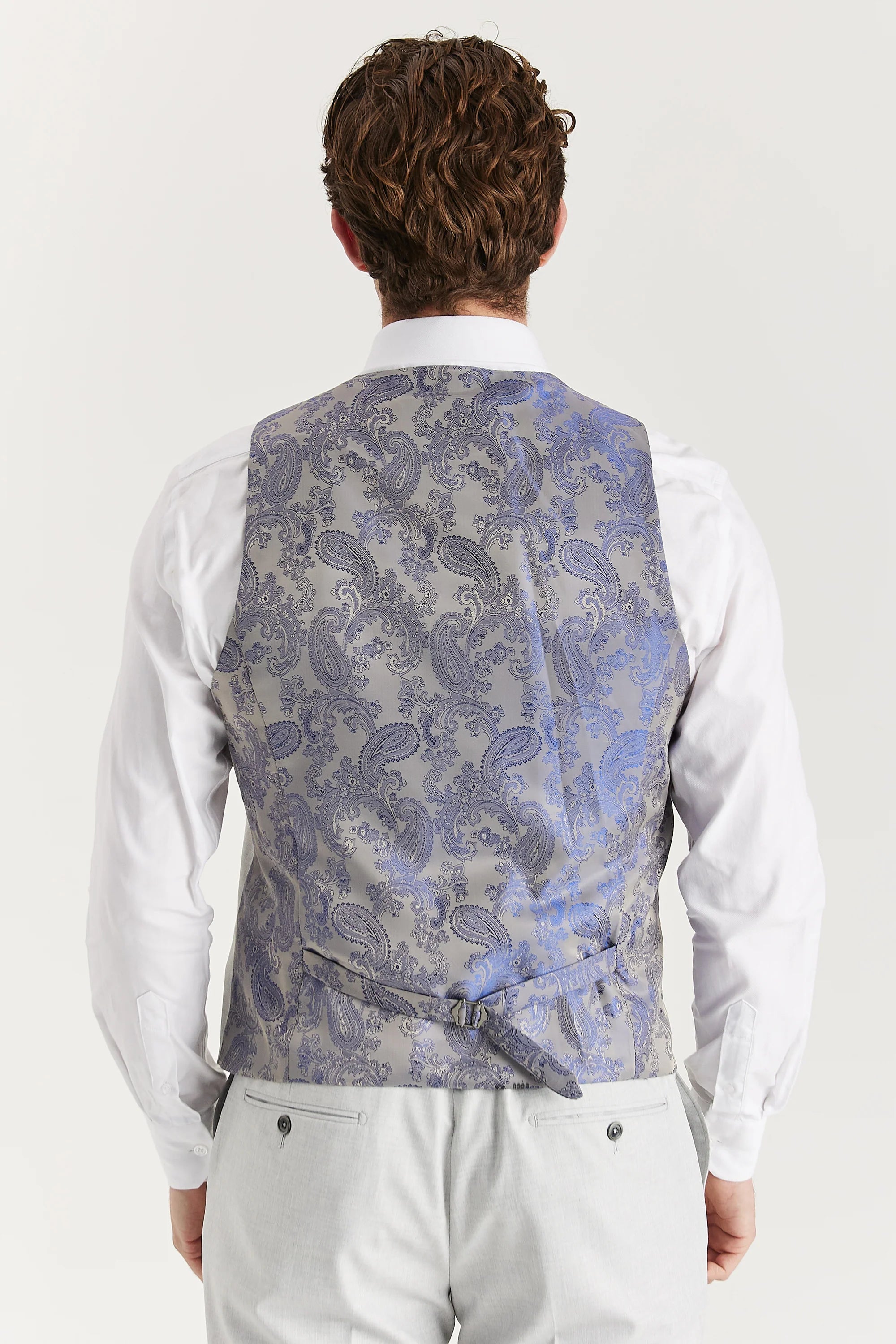 Rear view of the silver waistcoat with a silver paisley back panel and adjustable strap, worn with a white shirt and matching trousers.