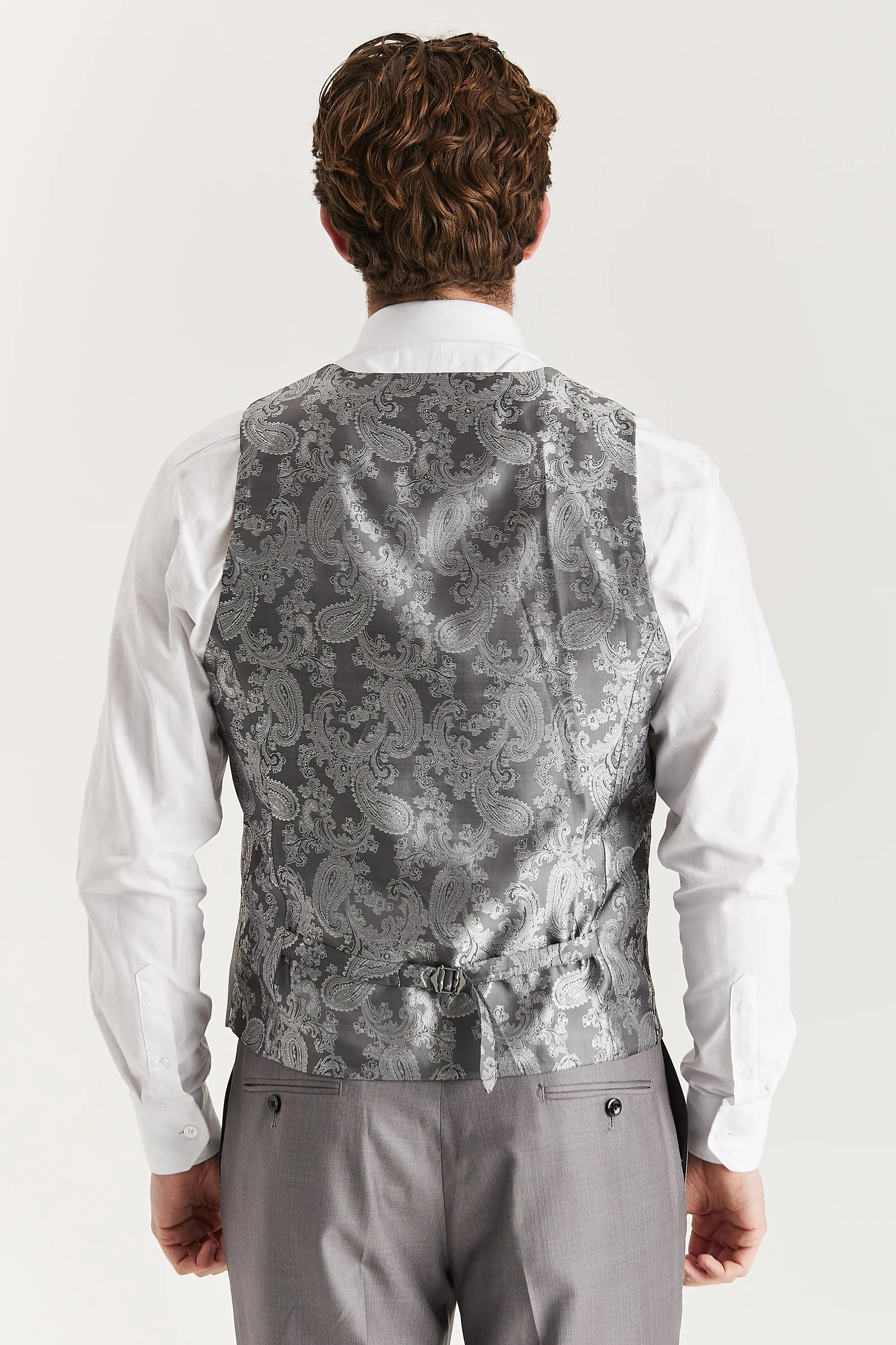 Rear view of the grey waistcoat with a silver paisley back panel and adjustable strap, worn with a white shirt and matching trousers.