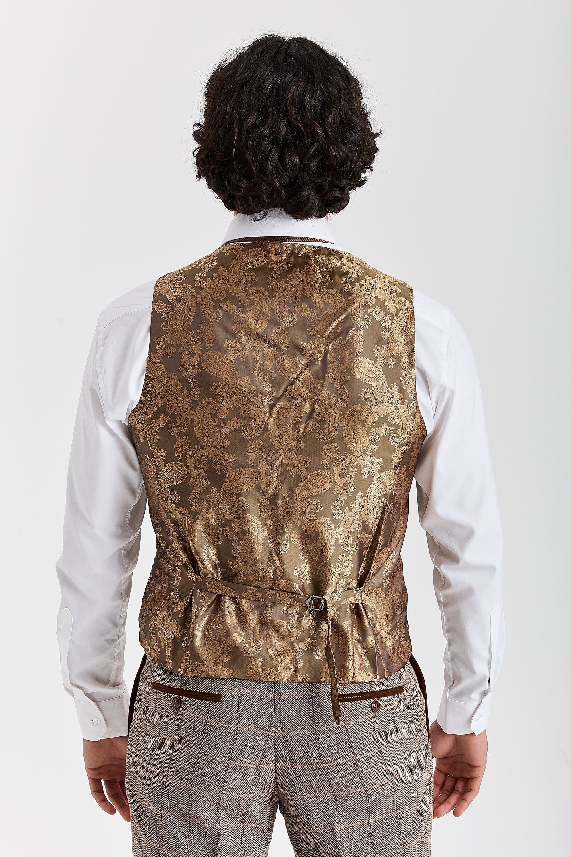 Back view of a golden brown paisley waistcoat panel with adjustable strap, worn with Harris brown herringbone tweed trousers and a white shirt.