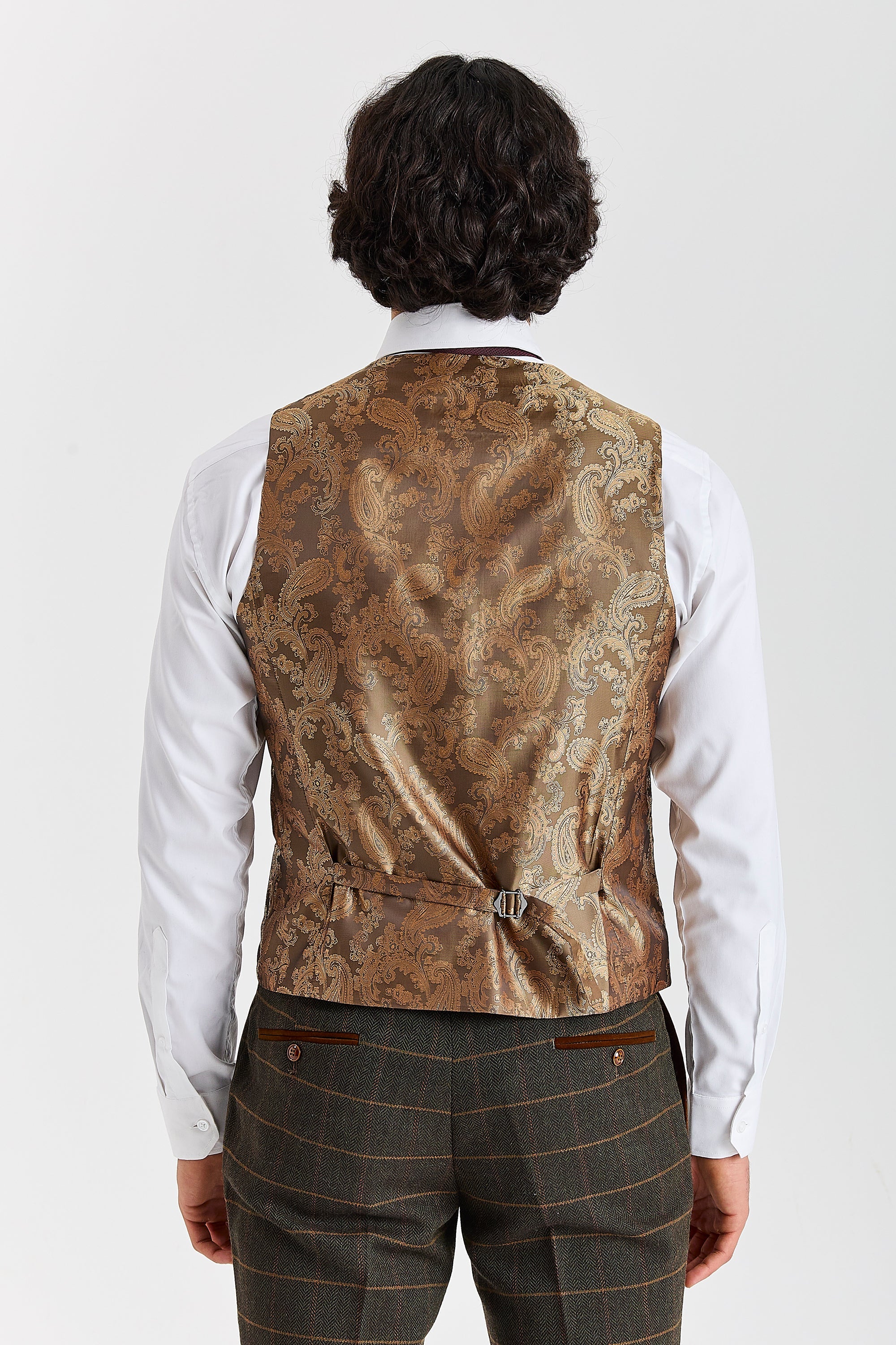 Back view of a waistcoat with gold paisley back panel and adjustable strap, worn with Harris green herringbone tweed check trousers and a white shirt.