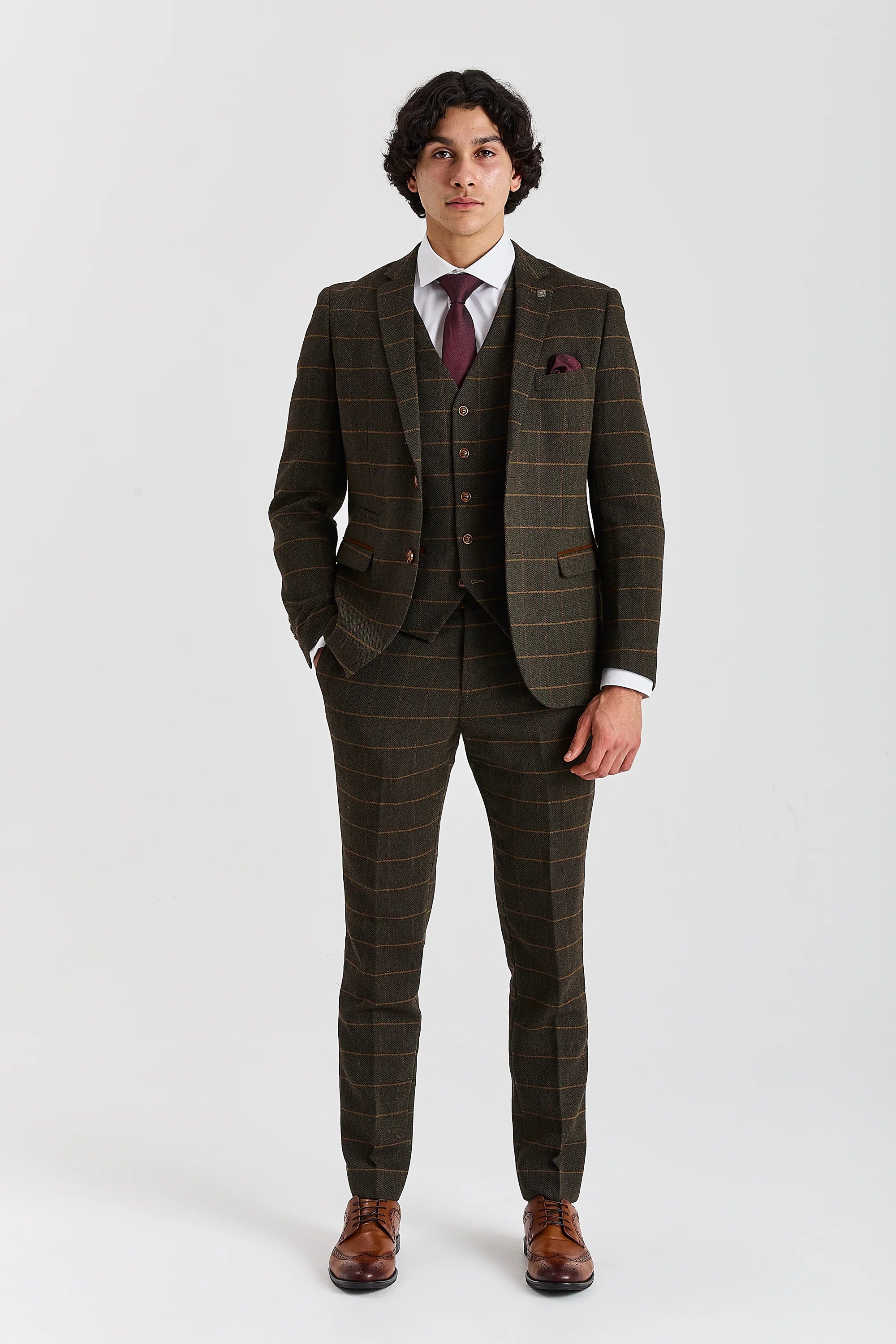 Full-length front view of a man wearing a Harris green herringbone tweed three piece suit with a tan windowpane check, burgundy tie and brown brogue shoes.