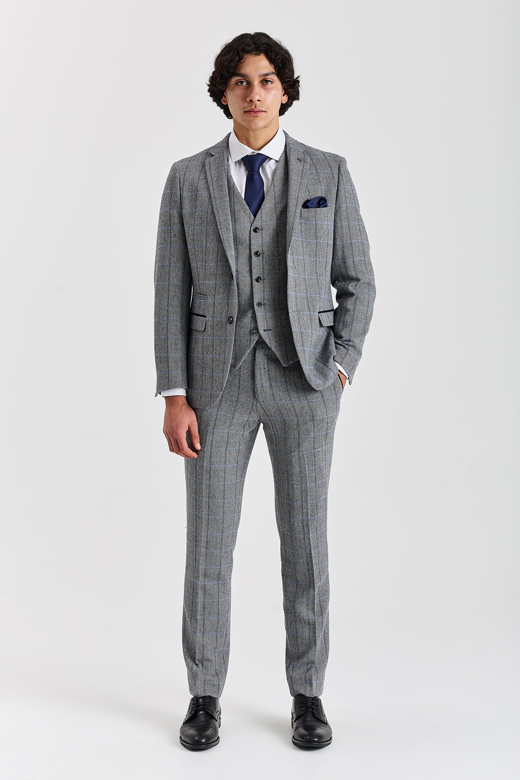 Full-length front view of a man wearing a Harris grey herringbone tweed check three piece suit with navy tie, pocket square and black shoes.