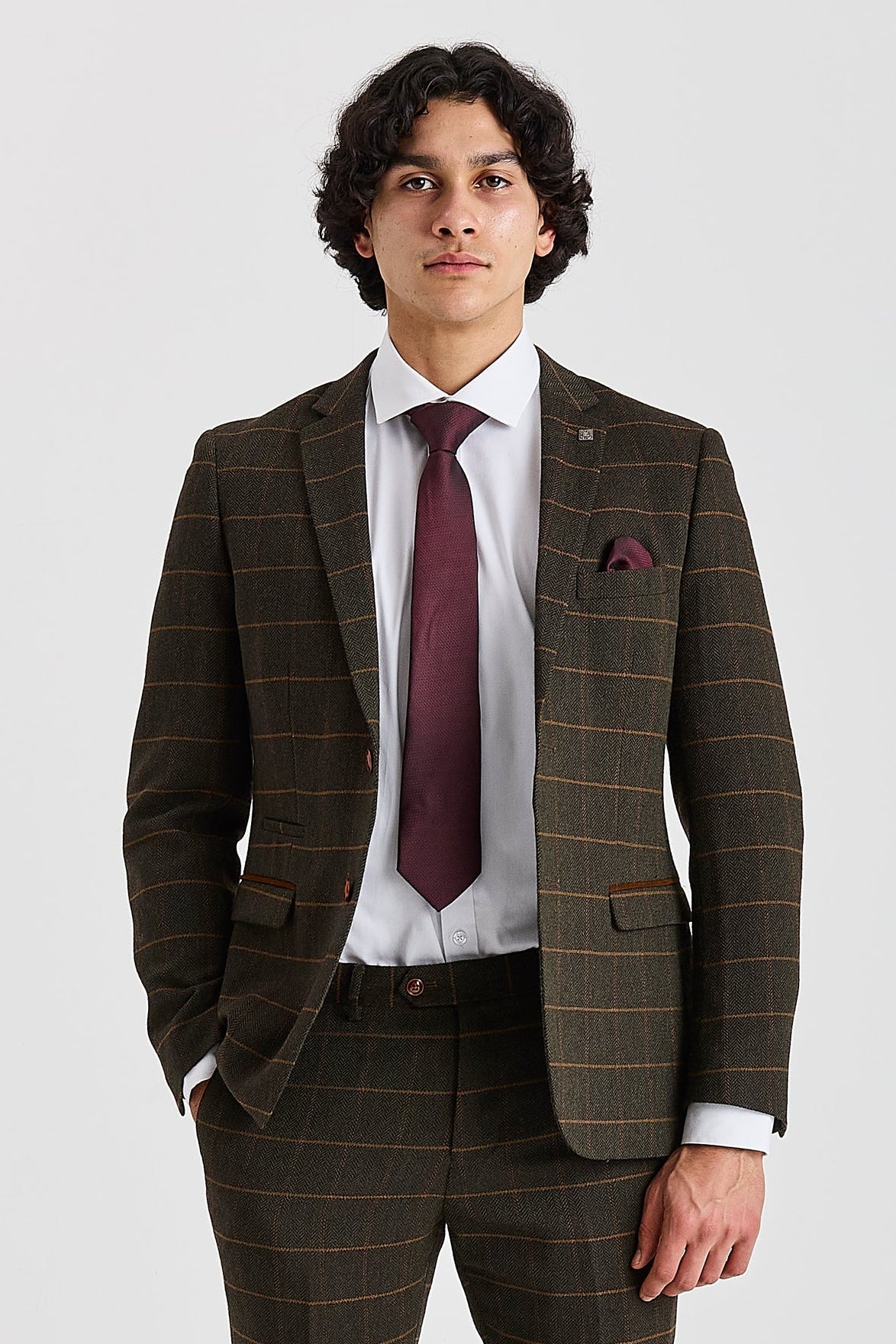 Knee-up front view of a man wearing a tailored green herringbone tweed suit jacket with a windowpane check pattern, styled with a crisp white shirt, burgundy tie, and matching burgundy pocket square.