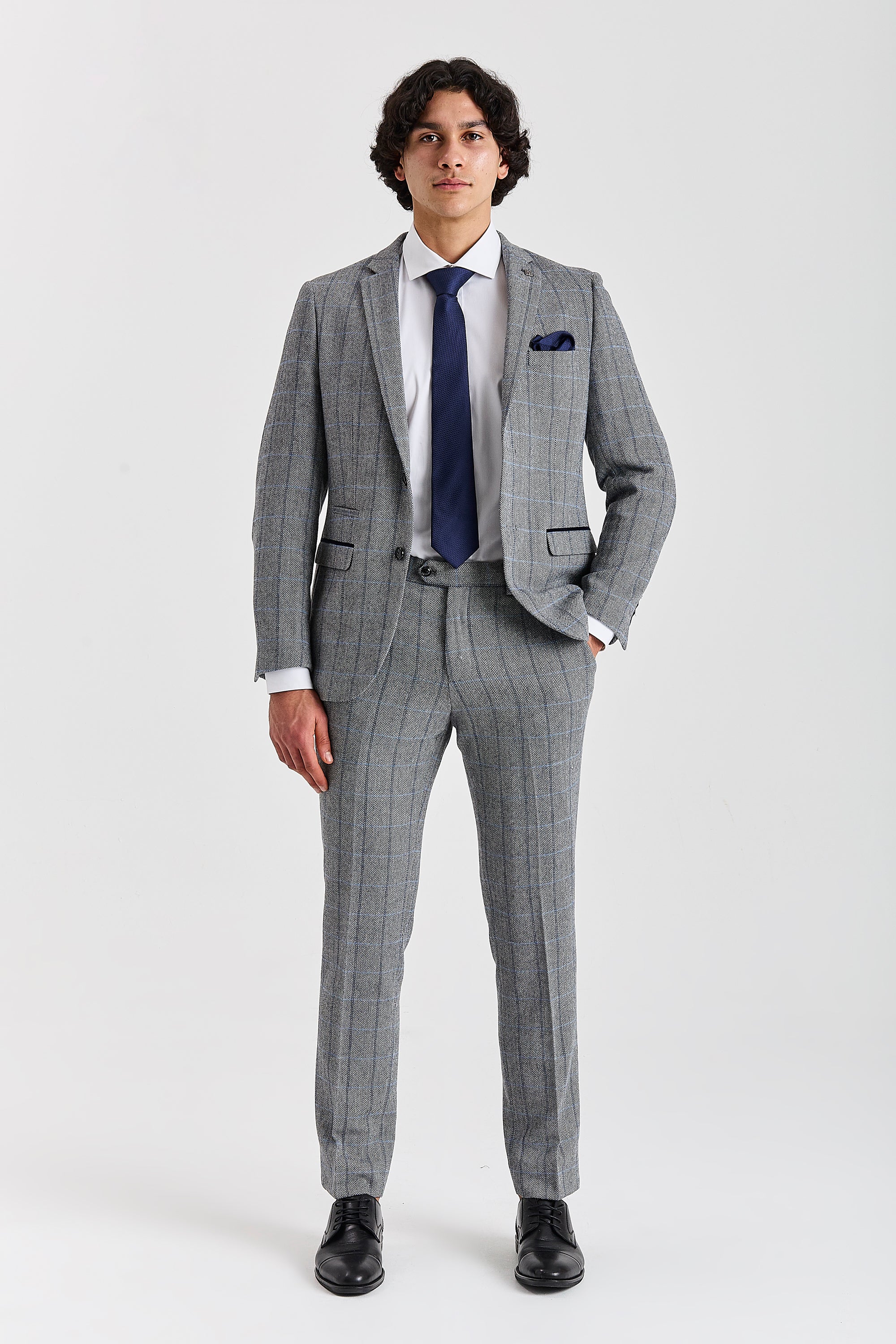 Full-length view of a man modeling a grey herringbone tweed two-piece suit with a windowpane check pattern. The outfit is completed with a white shirt, navy tie, and black leather shoes.