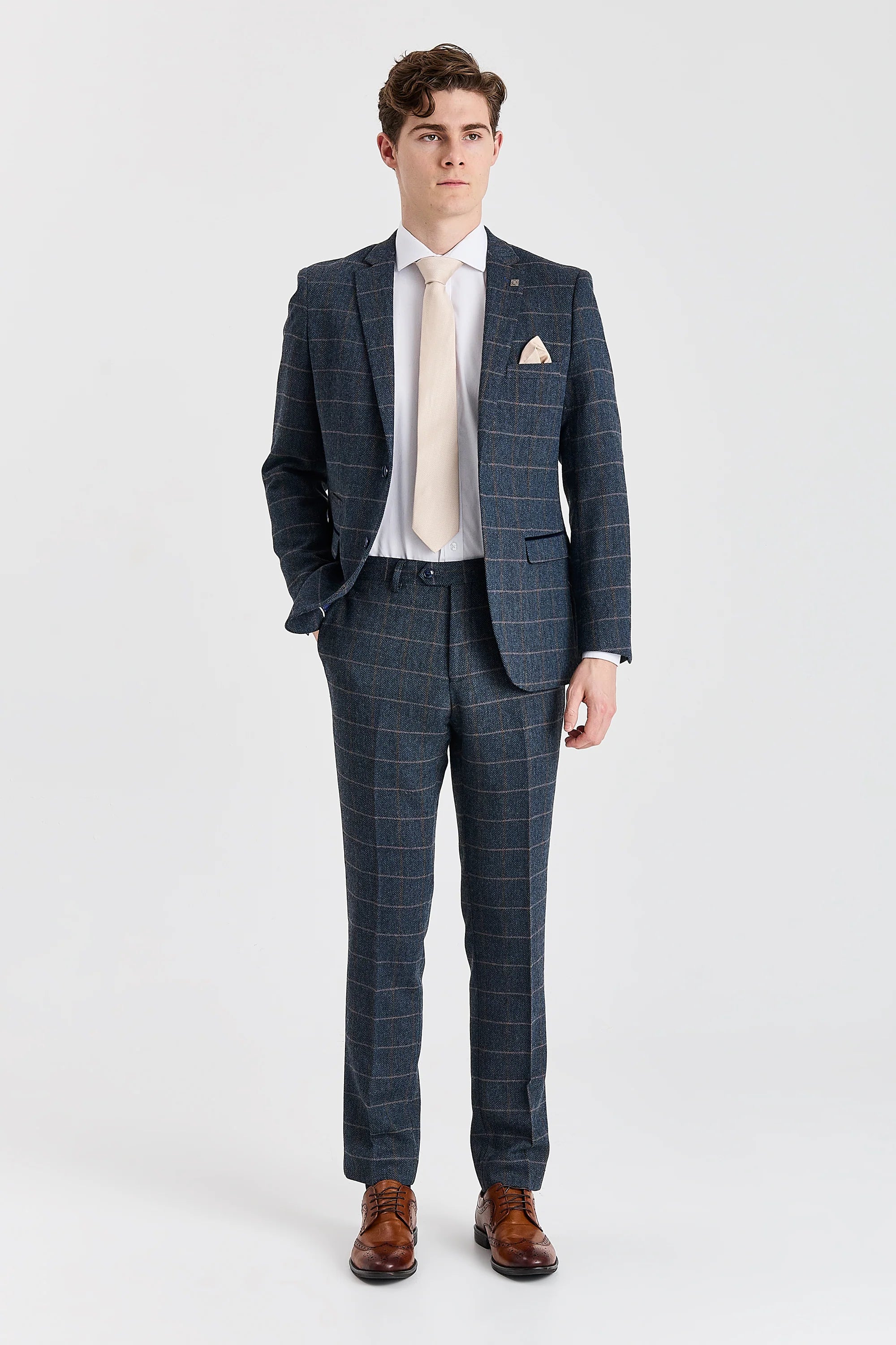 Full-length view of a man modeling a navy herringbone tweed two-piece suit with a windowpane check pattern. The outfit is completed with a white shirt, a light beige tie, and brown leather shoes.