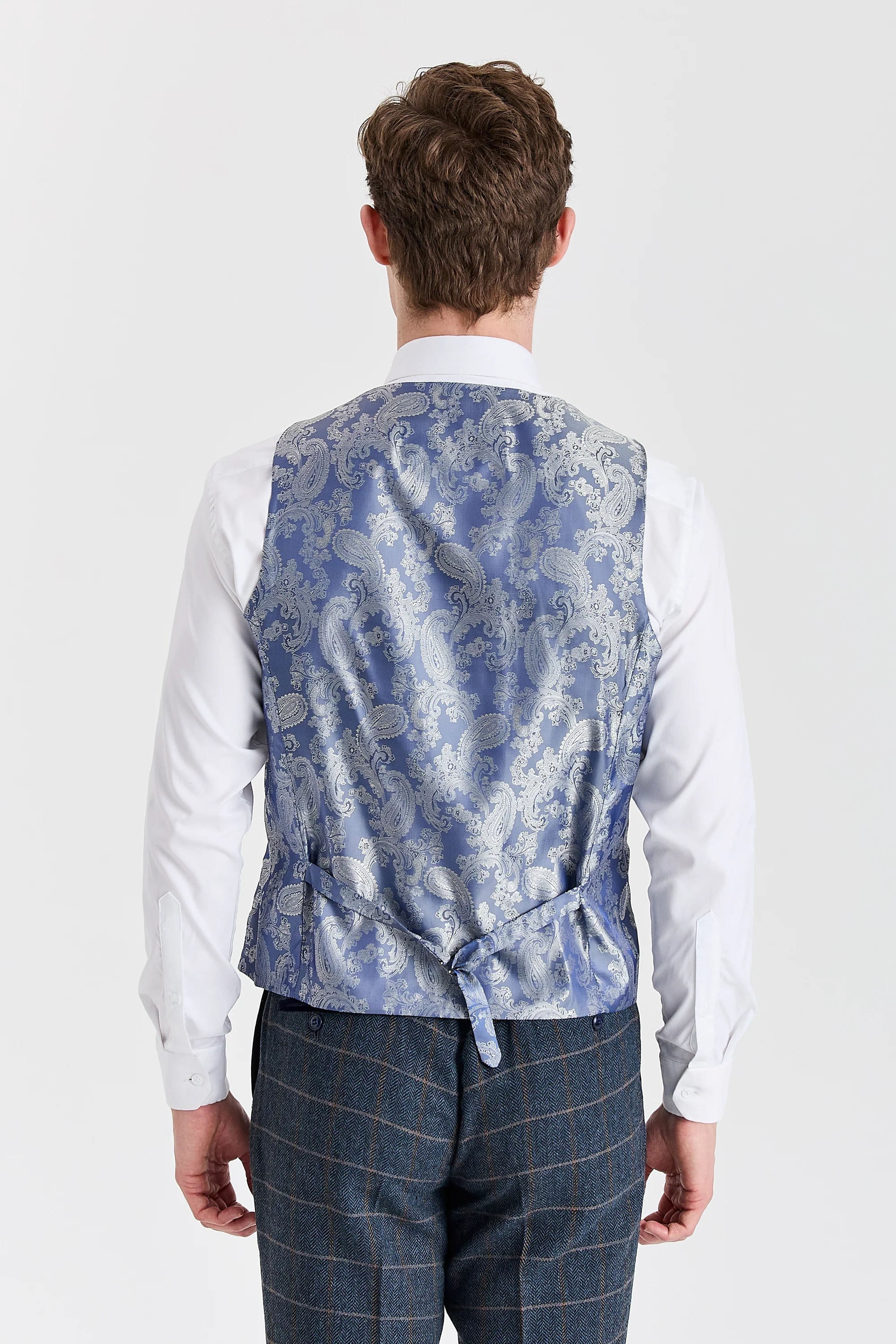 Back view of a waistcoat with blue and silver paisley back panel and adjustable strap, worn with Harris navy herringbone tweed check trousers and a white shirt.