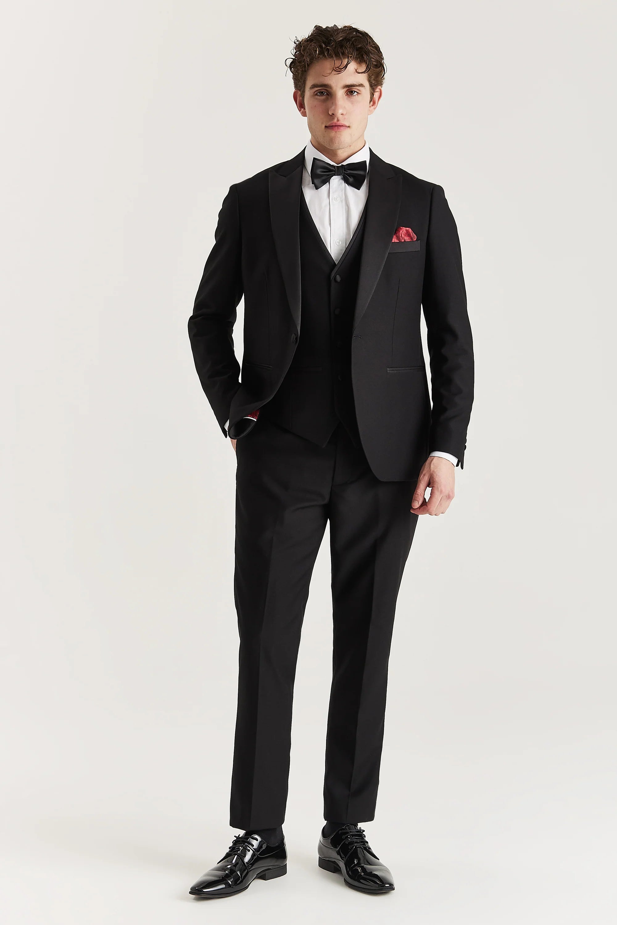 Full-length front view of a man wearing a black three piece dinner suit with satin peak lapels, waistcoat, trousers, white shirt, black bow tie, red pocket square and black shoes.