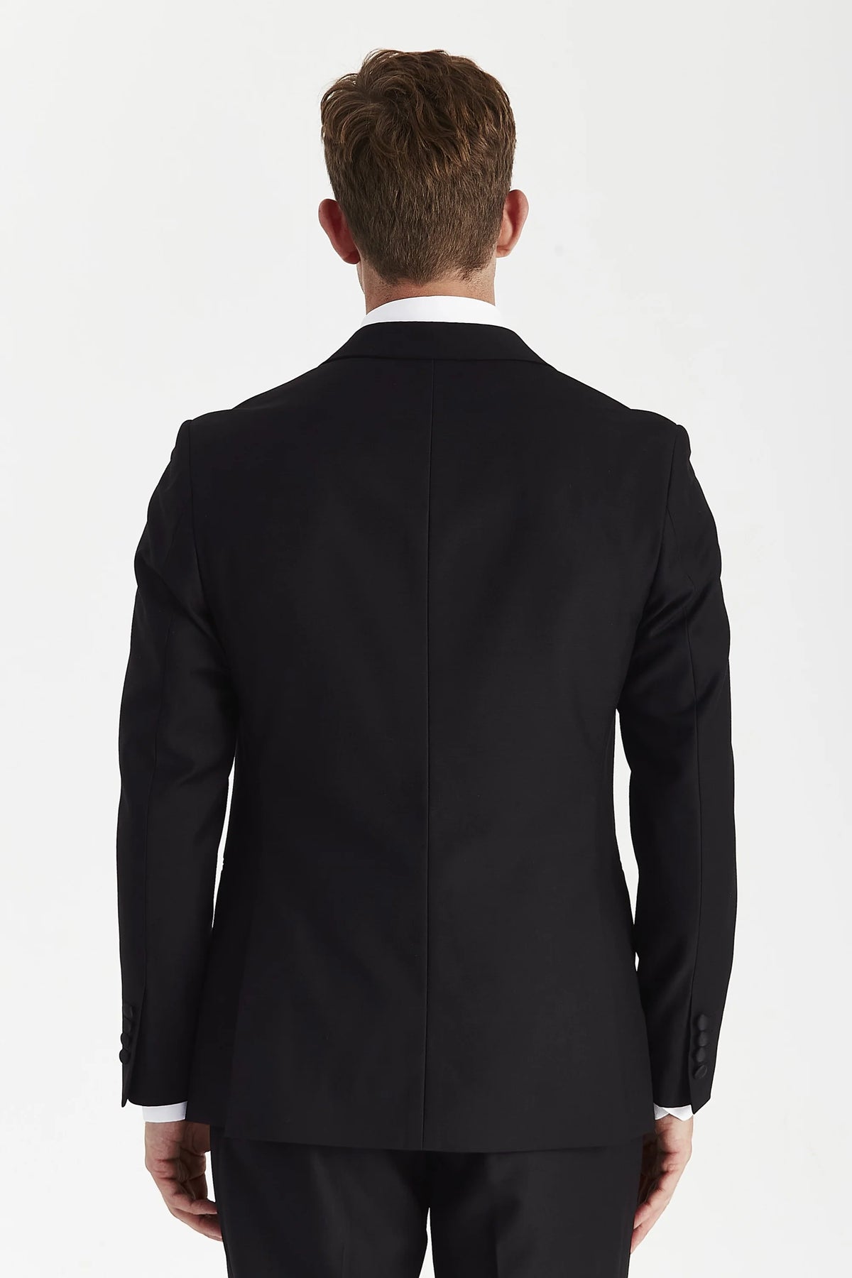 A man viewed from the back wearing a black suit jacket and matching trousers, with a white collared shirt visible at the neck, against a plain light background.